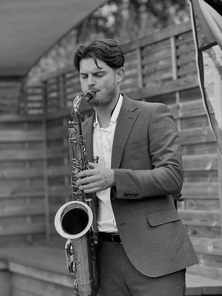 A young man in a suit playing a saxophone outdoors near a wooden fence.