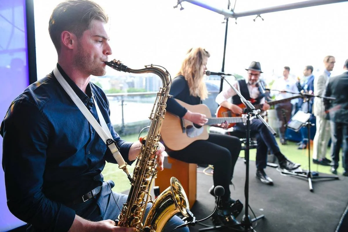 A band performing outdoors under a canopy, with a man playing the saxophone, a woman singing and playing guitar, and another man playing guitar, with several people in the background.