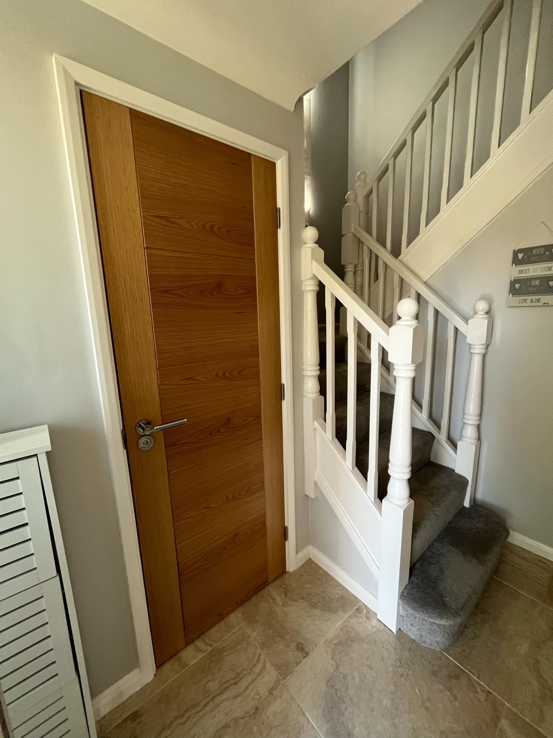 Indoors, wooden door, white staircase with railings, beige tiled floor, part of a white louvered door.