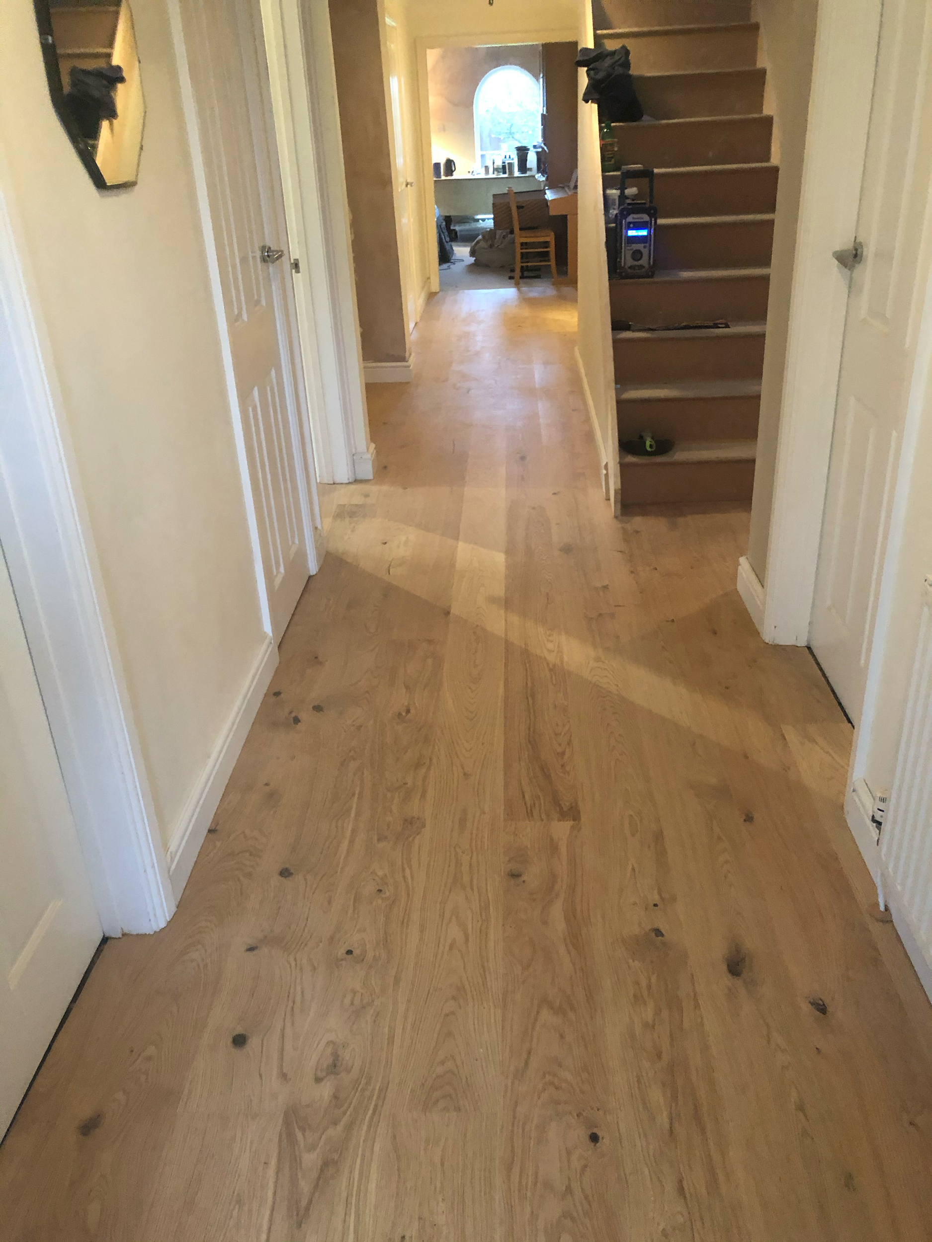 Interior hallway with wooden flooring, white paneled doors, staircase with visible wooden steps, and a glimpse into another room with a round window and furniture.