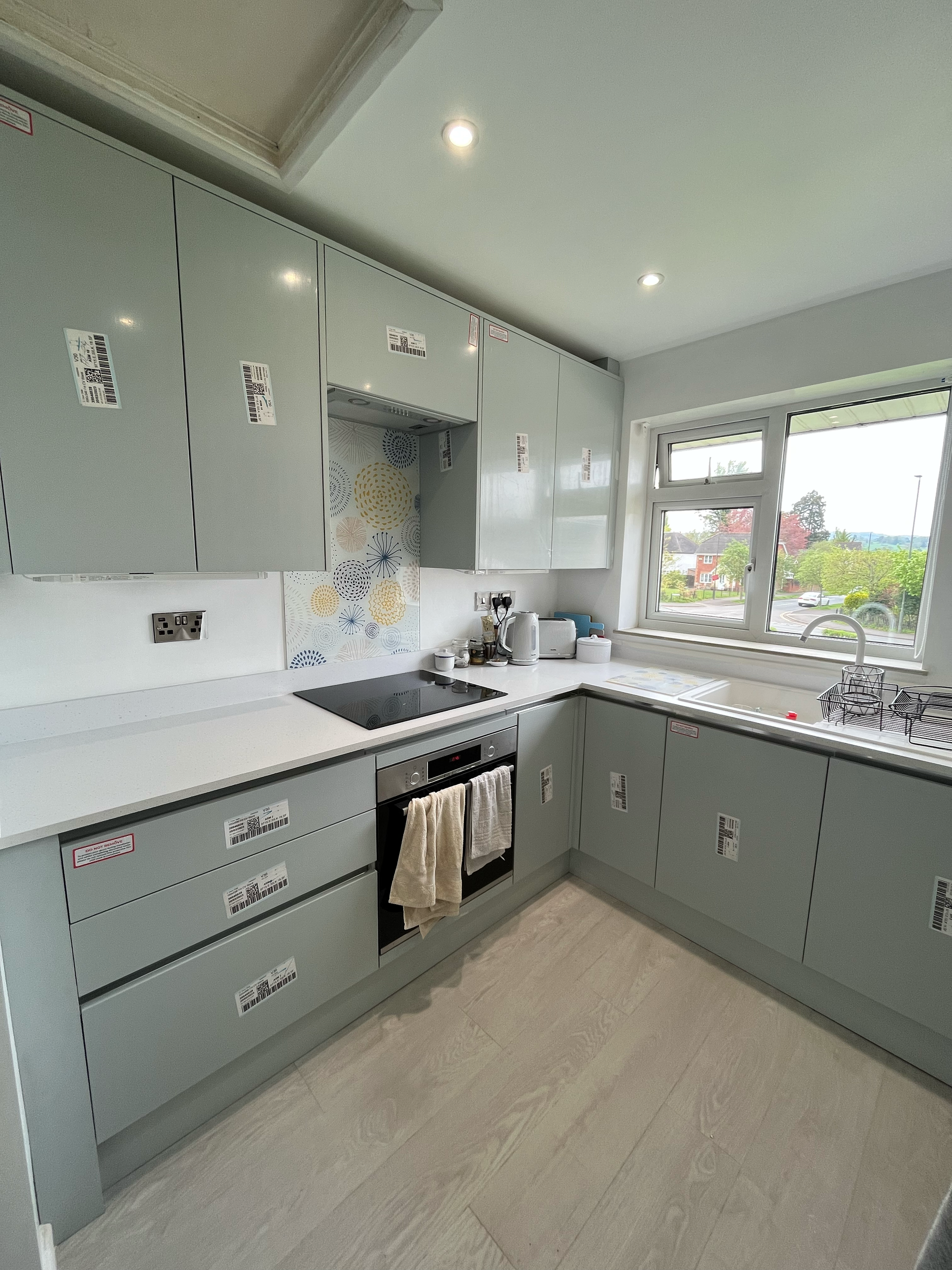 Modern kitchen interior with light gray cabinets, built-in oven, stovetop, sink, and a window overlooking a garden.
