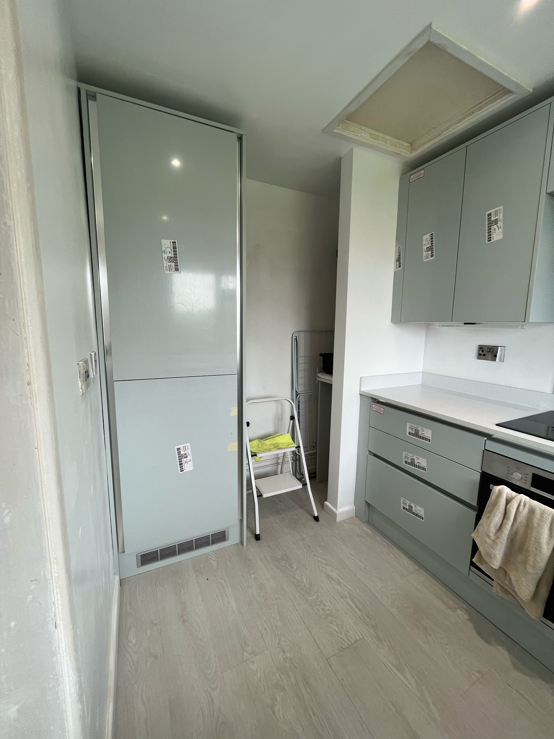 Modern kitchen with light gray cabinets, built-in oven, and refrigerator. Kitchen towel hanging on oven handle. Small step ladder and green chair against the wall. Bright, clean appearance.