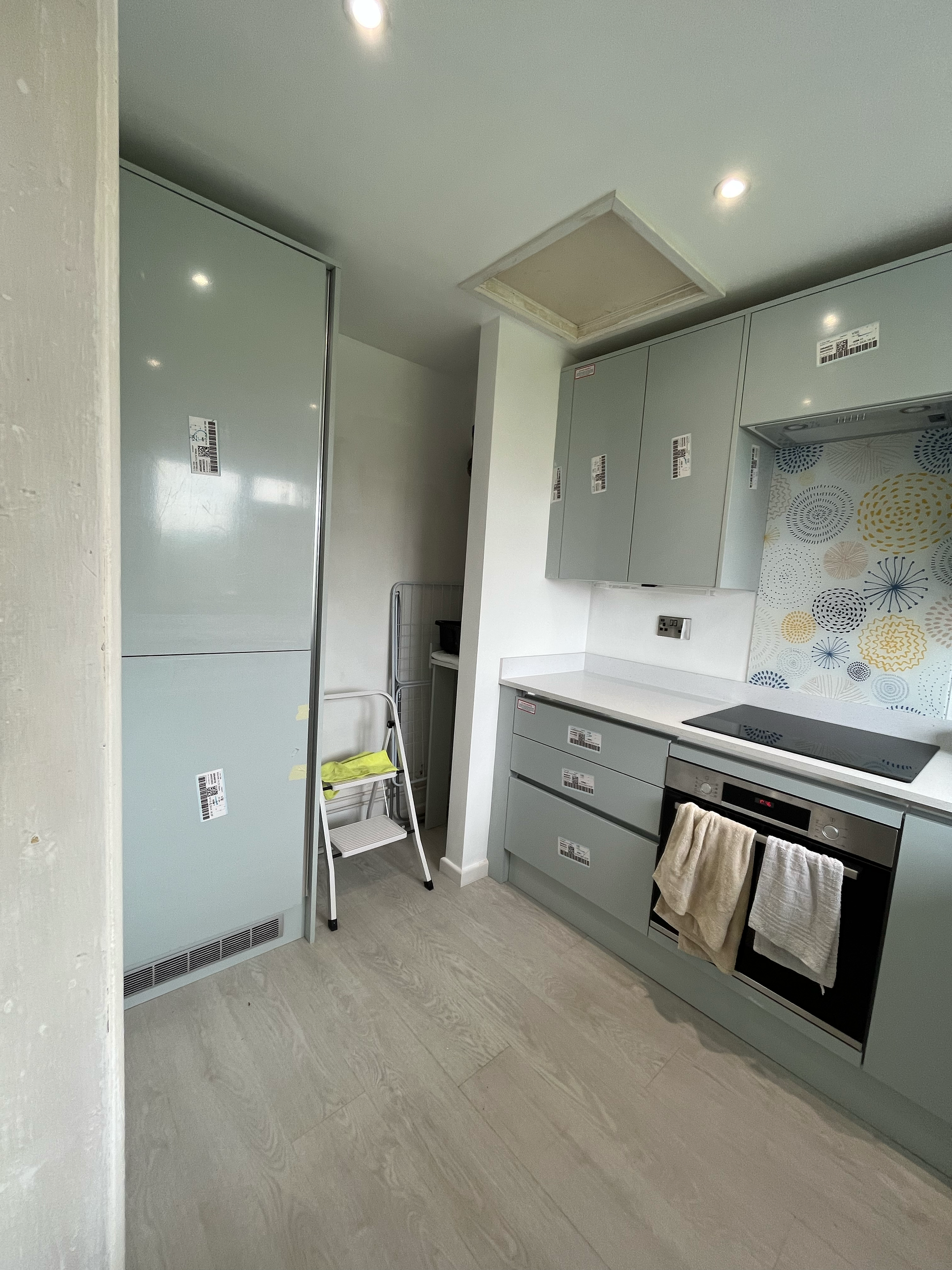 Modern kitchen with light gray cabinets, patterned backsplash, oven with towels hanging, and a small step ladder.