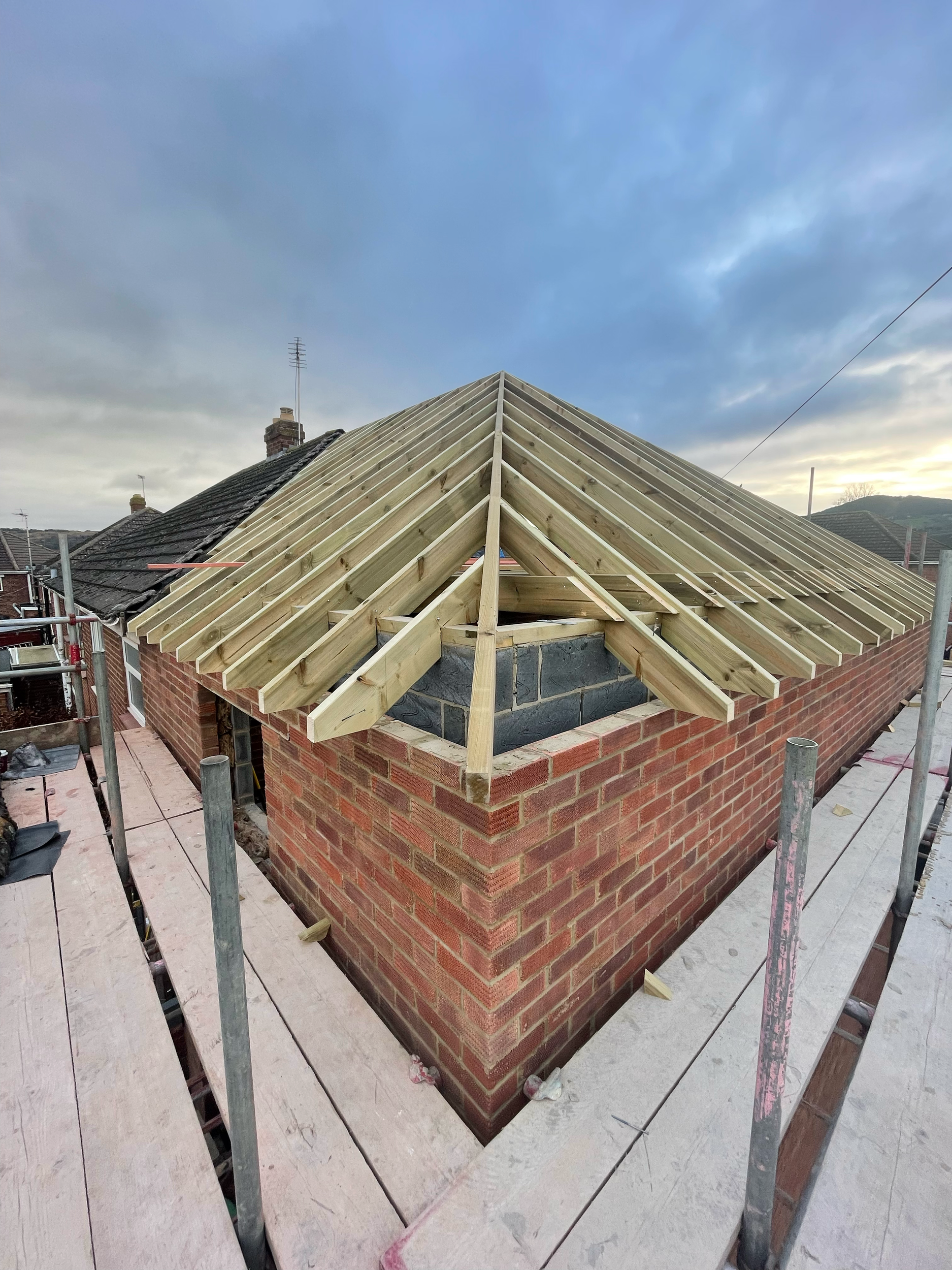 Construction site with wooden roof frame on a brick building, surrounded by scaffolding under a cloudy sky.