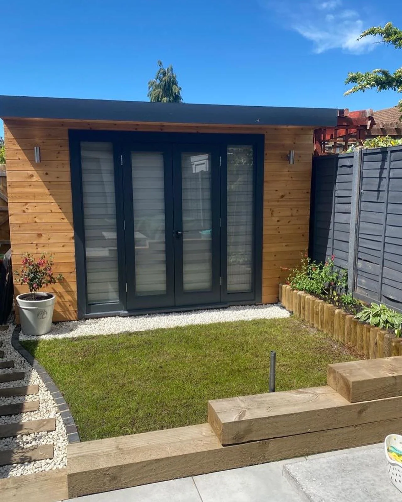 Modern wooden garden shed with glass doors, surrounded by grass, gravel, and a wooden fence. Small plants and shrubs are planted along the perimeter. Clear blue sky overhead.