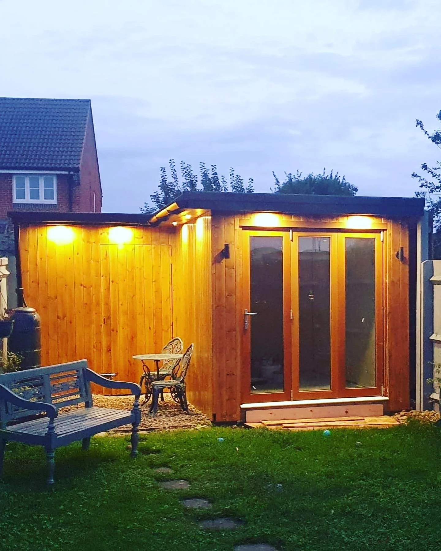 Wooden shed with glass doors and outdoor lights, a bench and metal chairs on a grassy yard, next to a brick house.