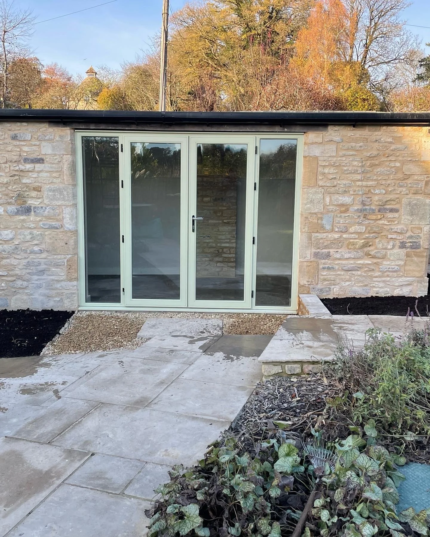 Stone building with large glass doors, surrounded by a paved area and garden, with trees in the background.