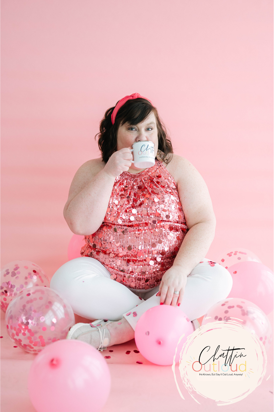 A woman sitting on the floor with pink and clear balloons around her, holding a white mug and wearing a pink sequined top and white pants, against a pink background.