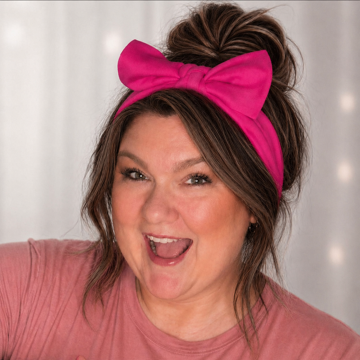 A woman with brown hair wearing a pink headband with a bow, smiling and making a playful face, against a light background.