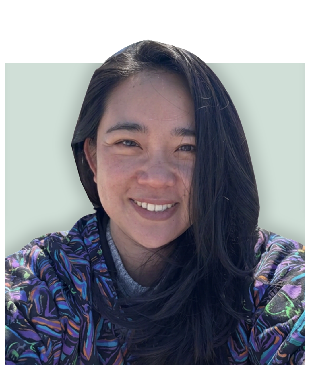 Close-up of a smiling woman with long dark hair, wearing a colorful patterned jacket, in front of a light-colored background.