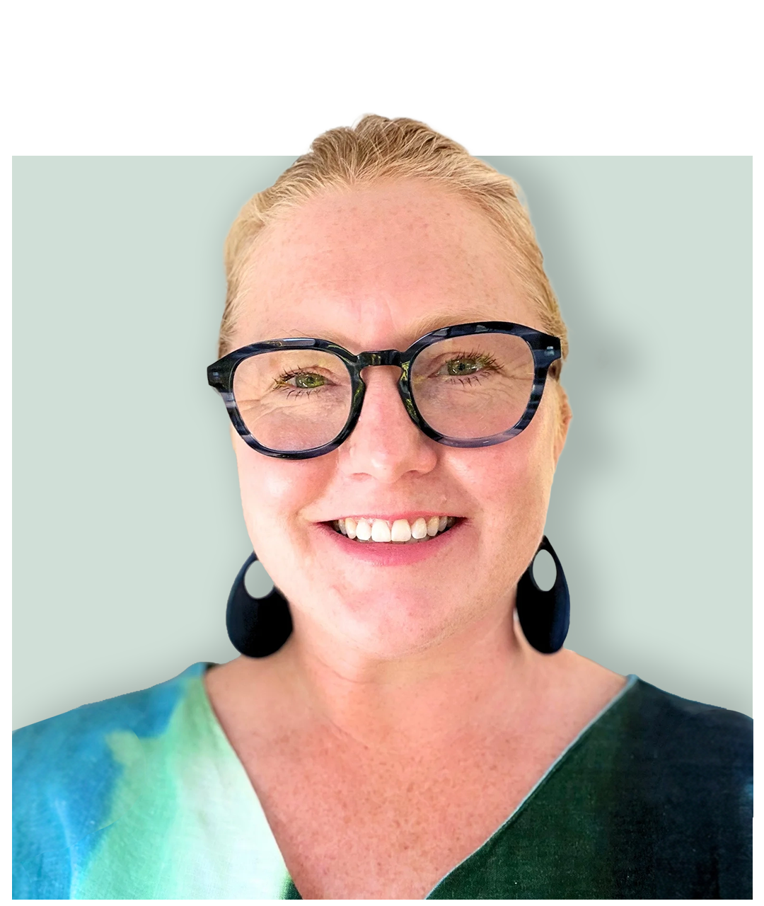 A woman with blonde hair, wearing large black glasses, black earrings, and a wet multicolored shirt, smiling against a light green background.