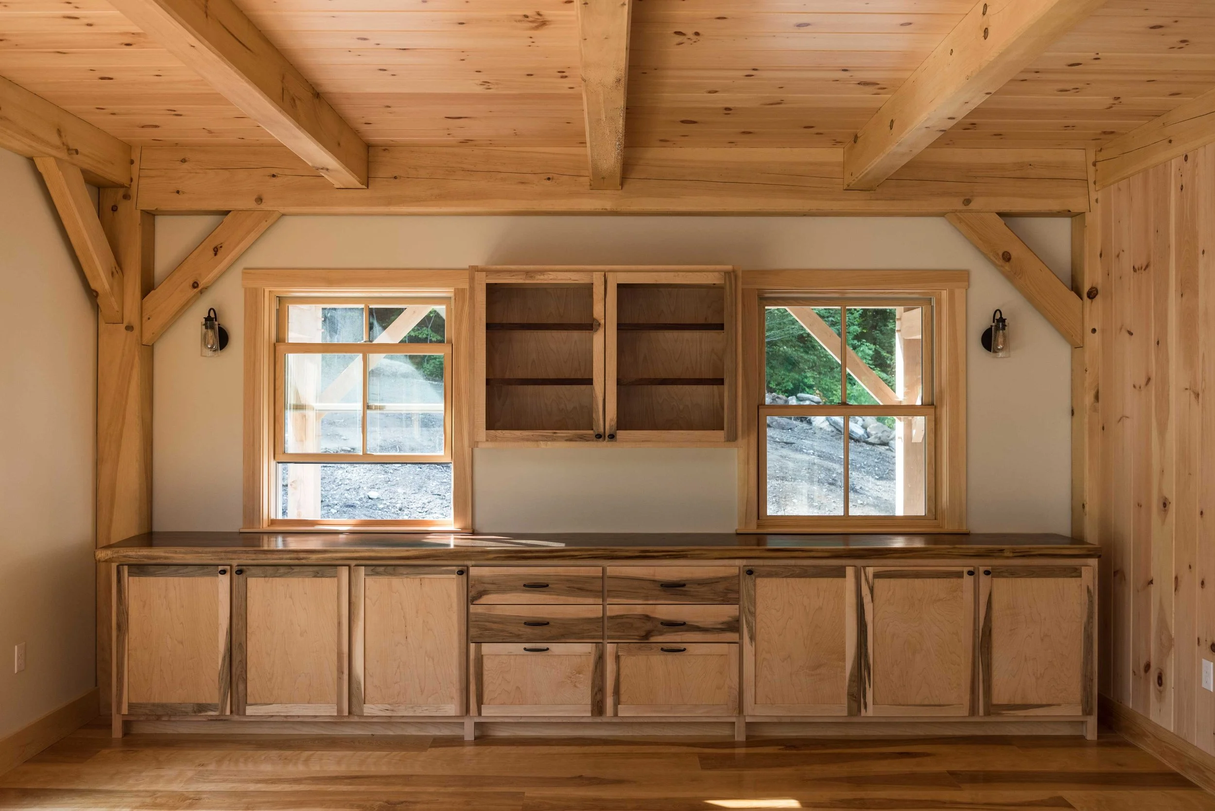 Interior of a rustic room with wooden cabinets, shelves, and exposed beams. The room features two windows and wood-paneled walls and ceiling.
