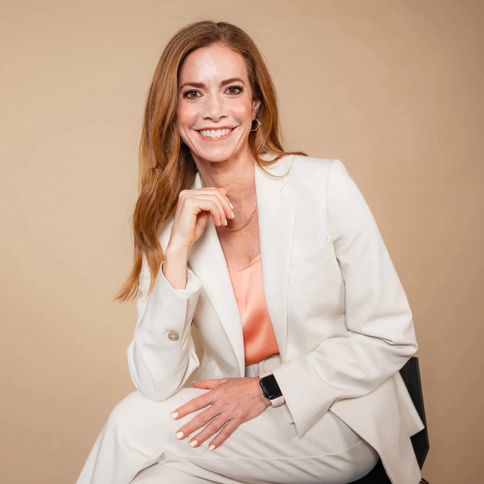 A woman with long red hair, smiling, sitting on a chair against a beige background. She is wearing a white blazer, a peach top, a watch, and a necklace.