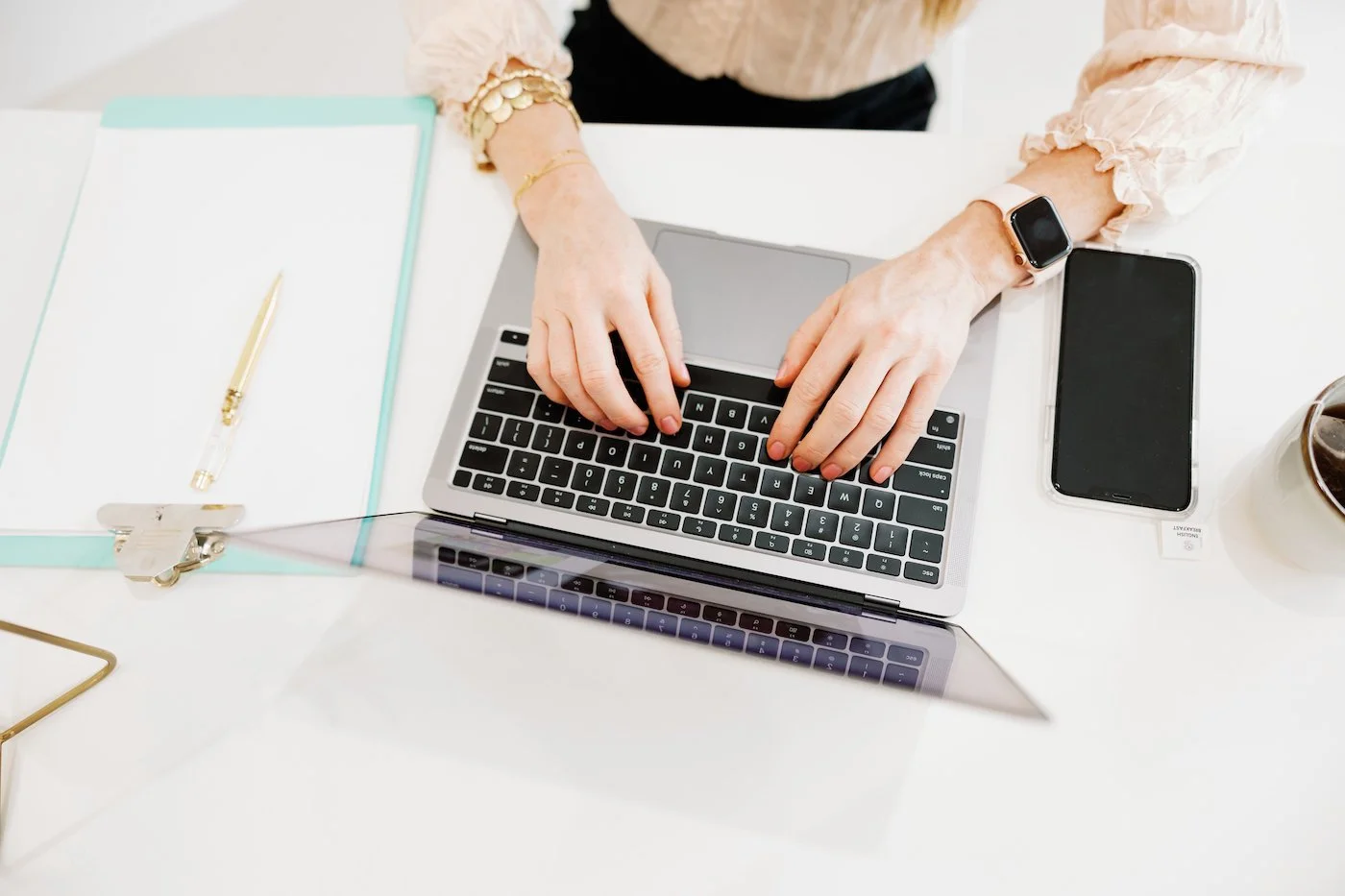 Person typing on a laptop at a white desk with a smartphone, a notebook, and a pen.