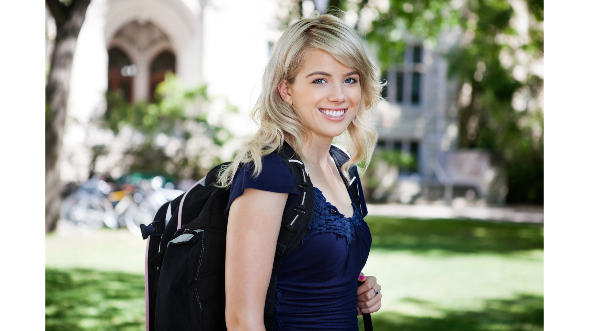 A young woman with blonde hair and blue eyes smiling outdoors, wearing a blue top and carrying a black backpack, with a park and buildings in the background.