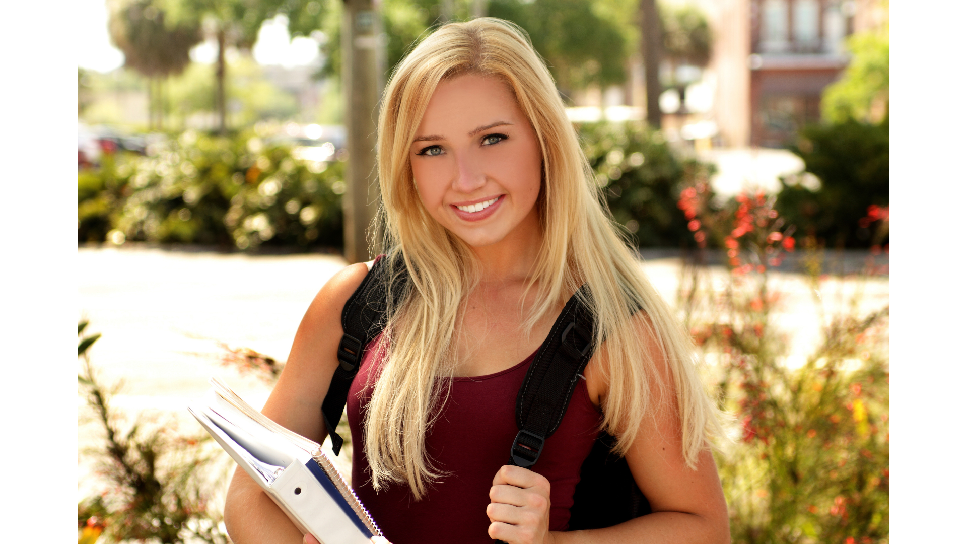 A young woman with long blonde hair, smiling, holding notebooks and wearing a black backpack, standing outdoors in a sunny park or campus setting.