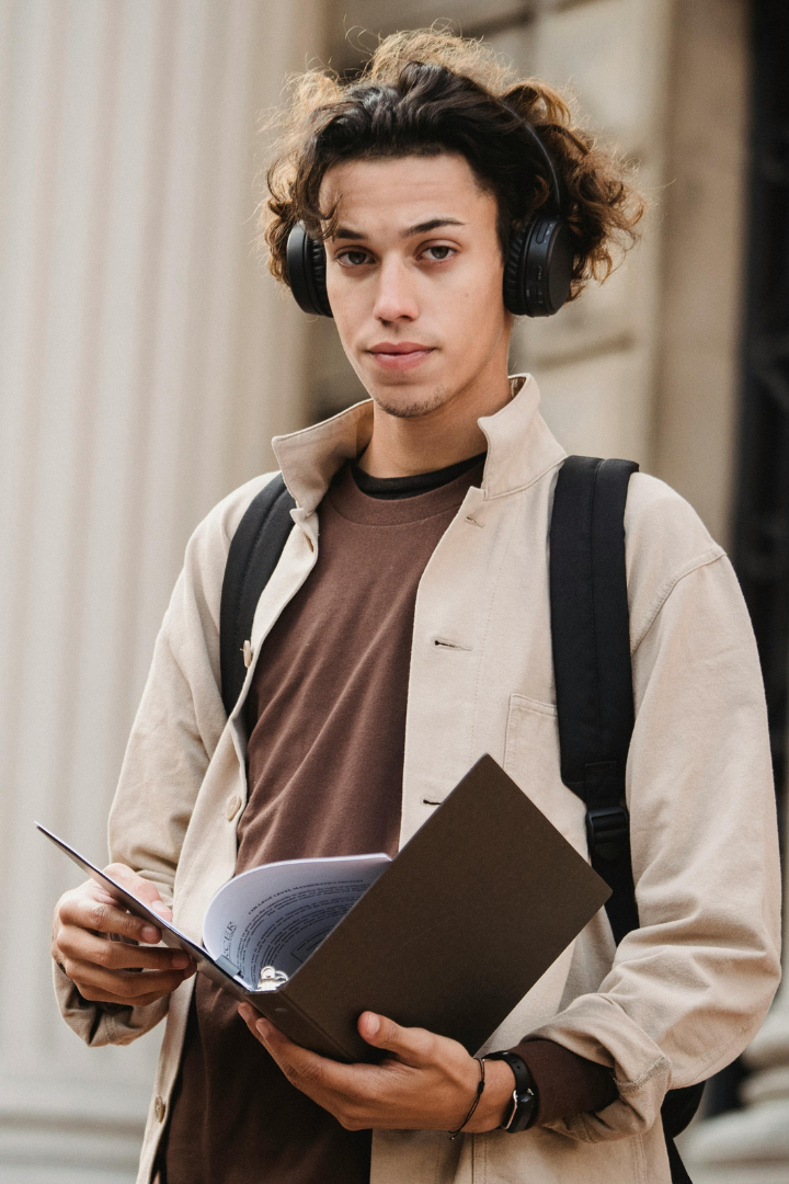 Young man with curly hair wearing headphones, holding an open book and a smartphone, with a backpack on his shoulder.