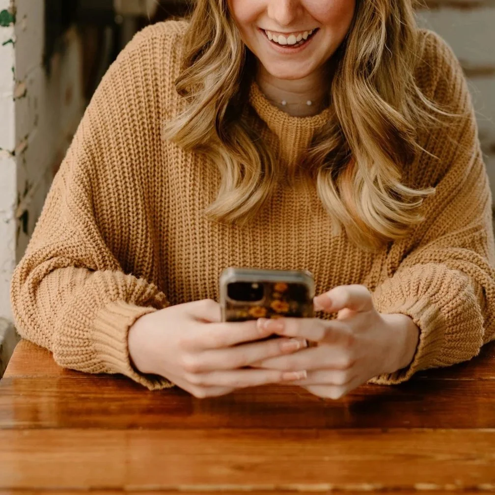 A woman with long wavy blonde hair smiling while looking at her phone, wearing a tan knitted sweater, sitting at a wooden table.