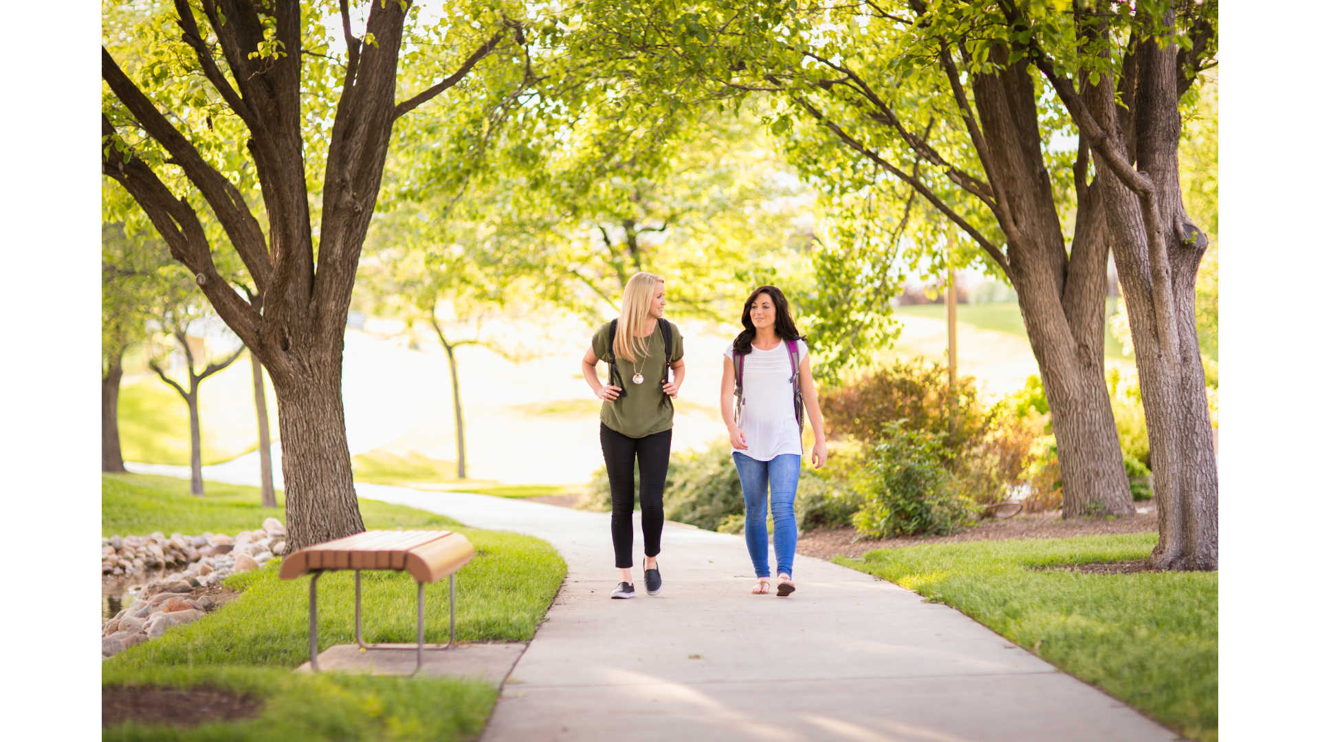 Two women walking and talking in a park on a sunny day, surrounded by green trees and grass.
