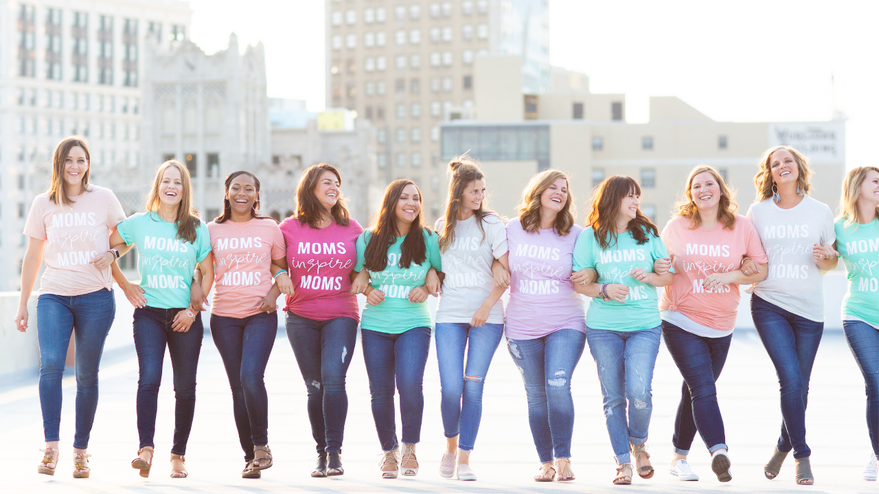 Group of women in colorful t-shirts that say 'Moms Inspire Moms' standing on a rooftop, linked arm-in-arm and smiling, with city buildings in the background.