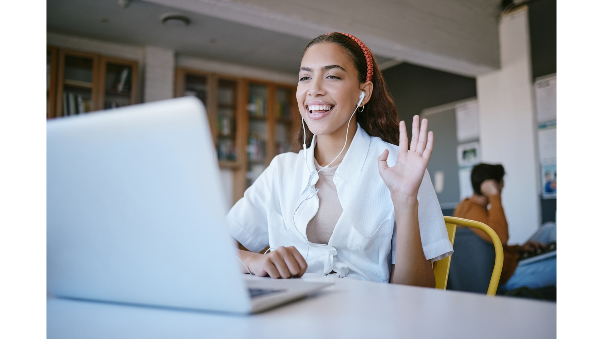 A young woman with brown hair wearing a red headband and white shirt, smiling and waving while participating in a video call on her laptop, with earphones in her ears, in a library or study area.