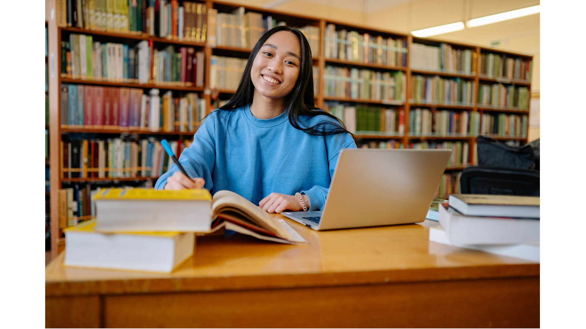 A young woman sitting at a wooden table in a library, smiling, with books, an open notebook, a laptop, and a pen in her hands.