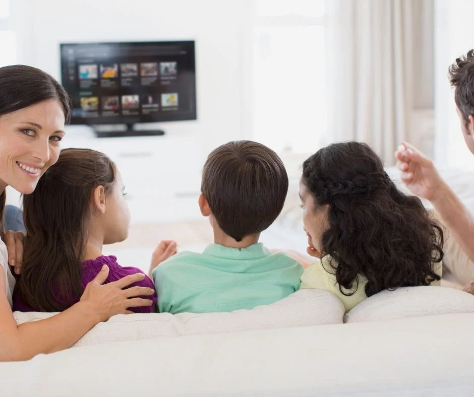 Family watching TV together in living room, seen from behind, with a woman turning and smiling at the camera.