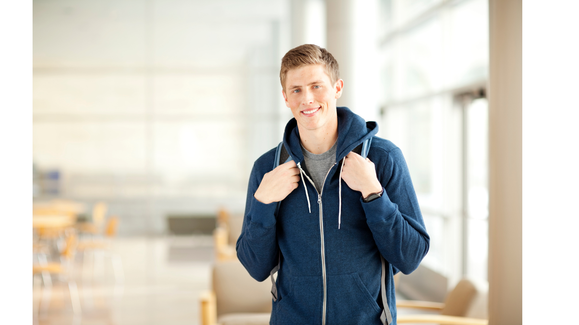 Young man wearing a blue hoodie with a backpack, standing inside a bright modern building, smiling at the camera.
