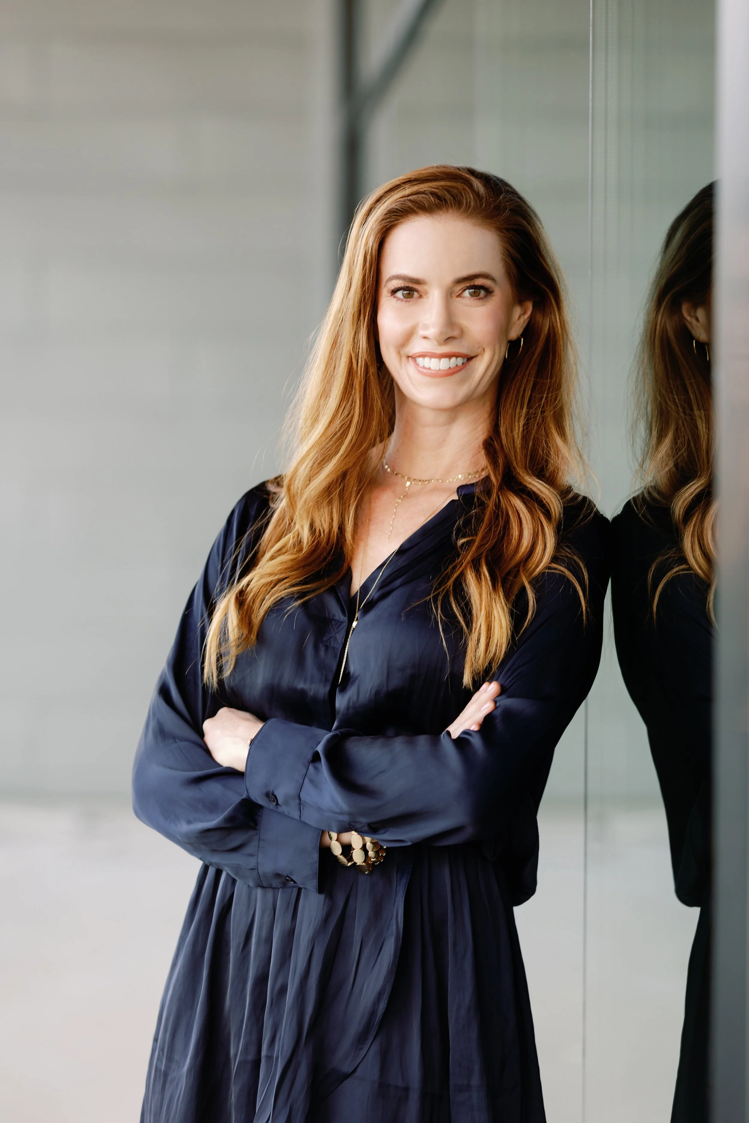 A smiling woman with long red hair standing with her arms crossed, leaning against a reflective glass wall.