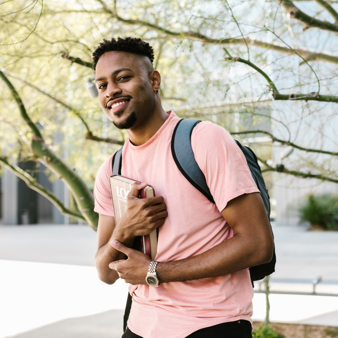 A smiling young man with a backpack holding a notebook outdoors near trees.