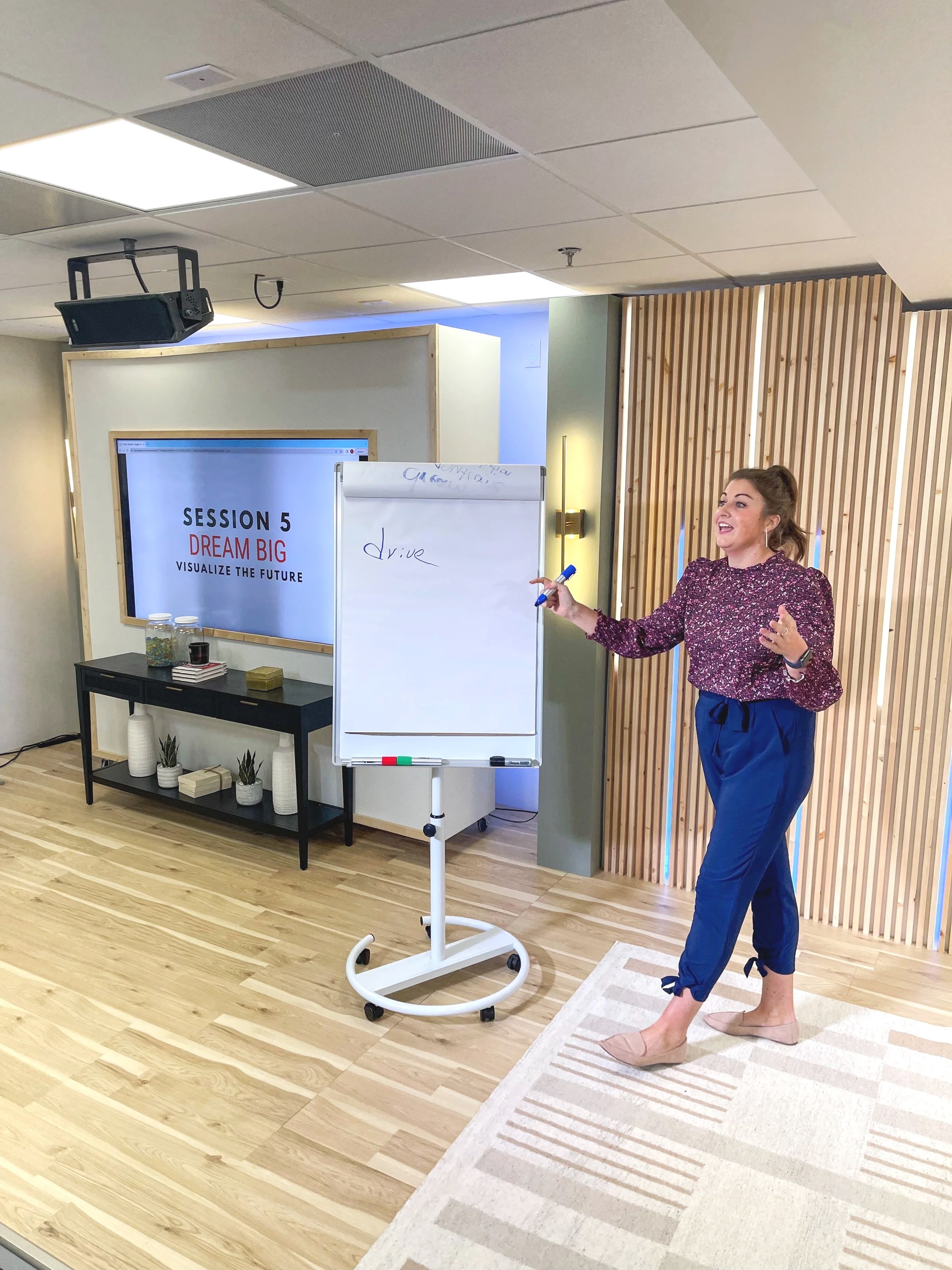 A woman giving a presentation in a conference room, standing barefoot on a rug, wearing a purple blouse and blue pants, with a whiteboard, a TV screen that says 'Session 5 Dream Big Visualize the Future,' and a table with decorative vases and books.
