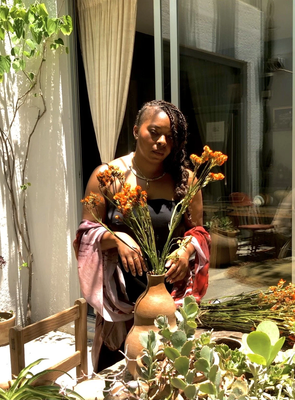 A woman standing outdoors at a table with potted plants and a large vase with orange flowers. She has dark curly hair, wears a black top, and a pink and red shawl. The background includes a sliding glass door and a balcony.