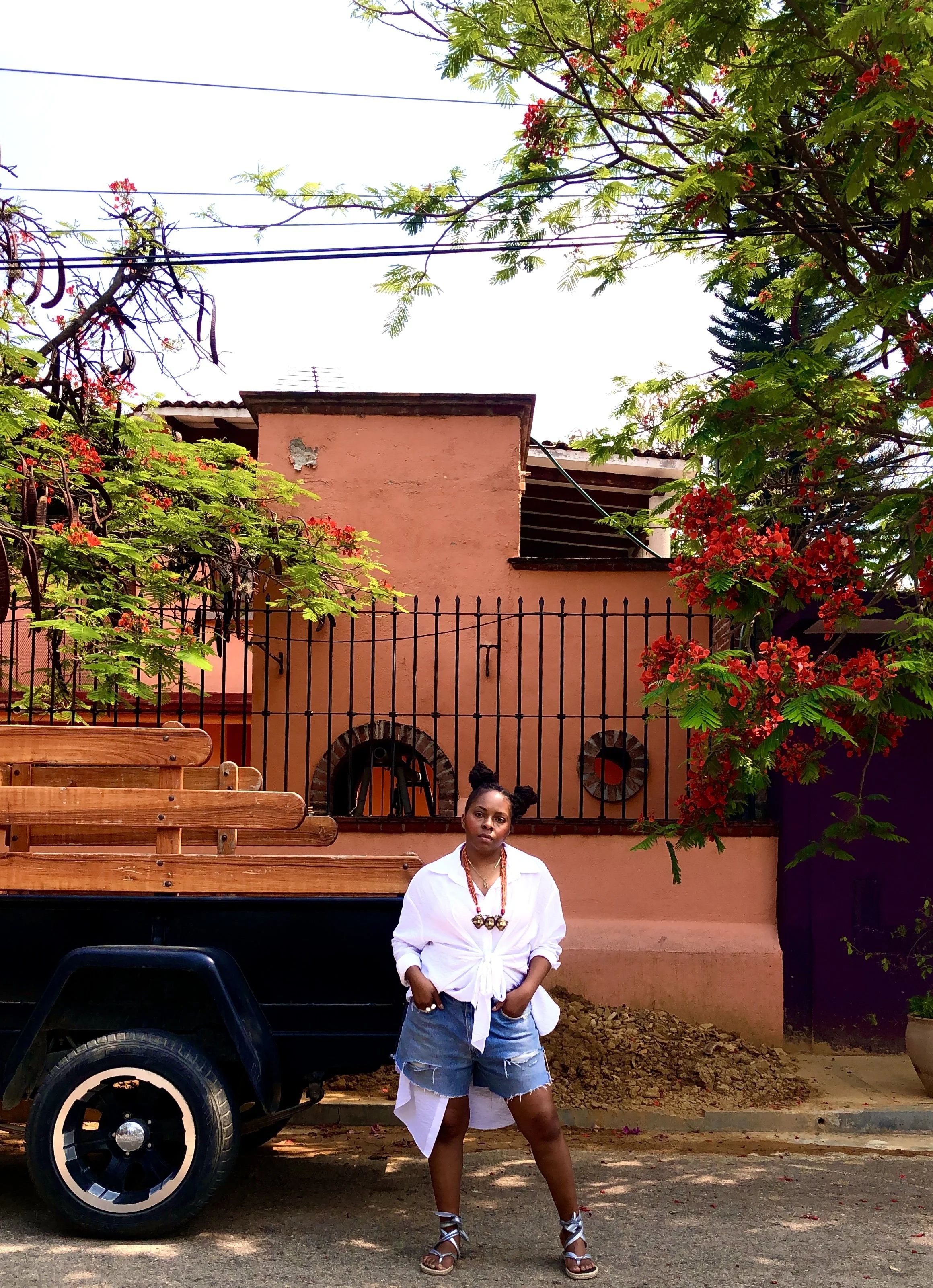 A woman standing on a street in front of a pink building with flowering trees and a black fence. She wears a white shirt, distressed denim shorts, and sandals, with her hands in her pockets and an expression of confidence.