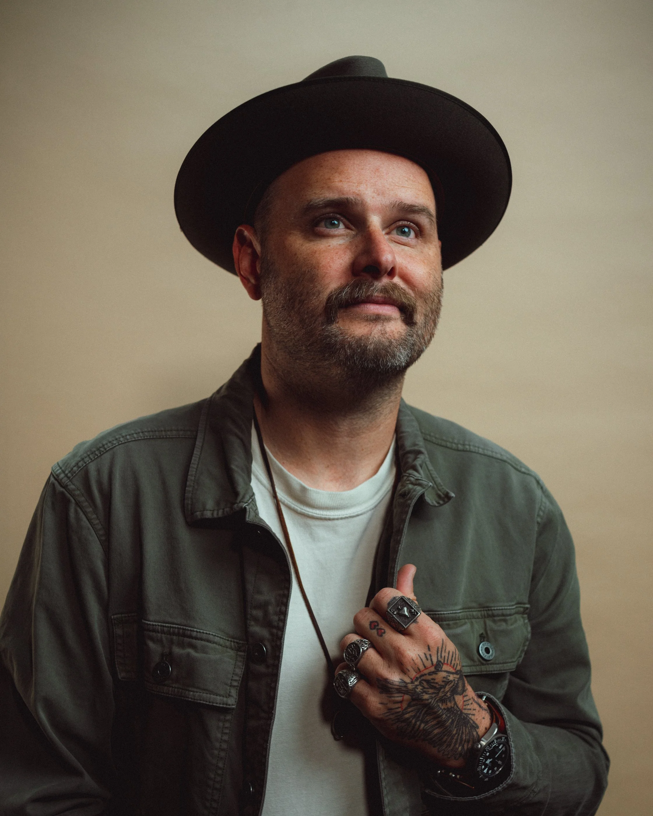 Dave Herring in a dark hat and olive overshirt looking up, one tattooed hand on his chest, against a warm tan backdrop.