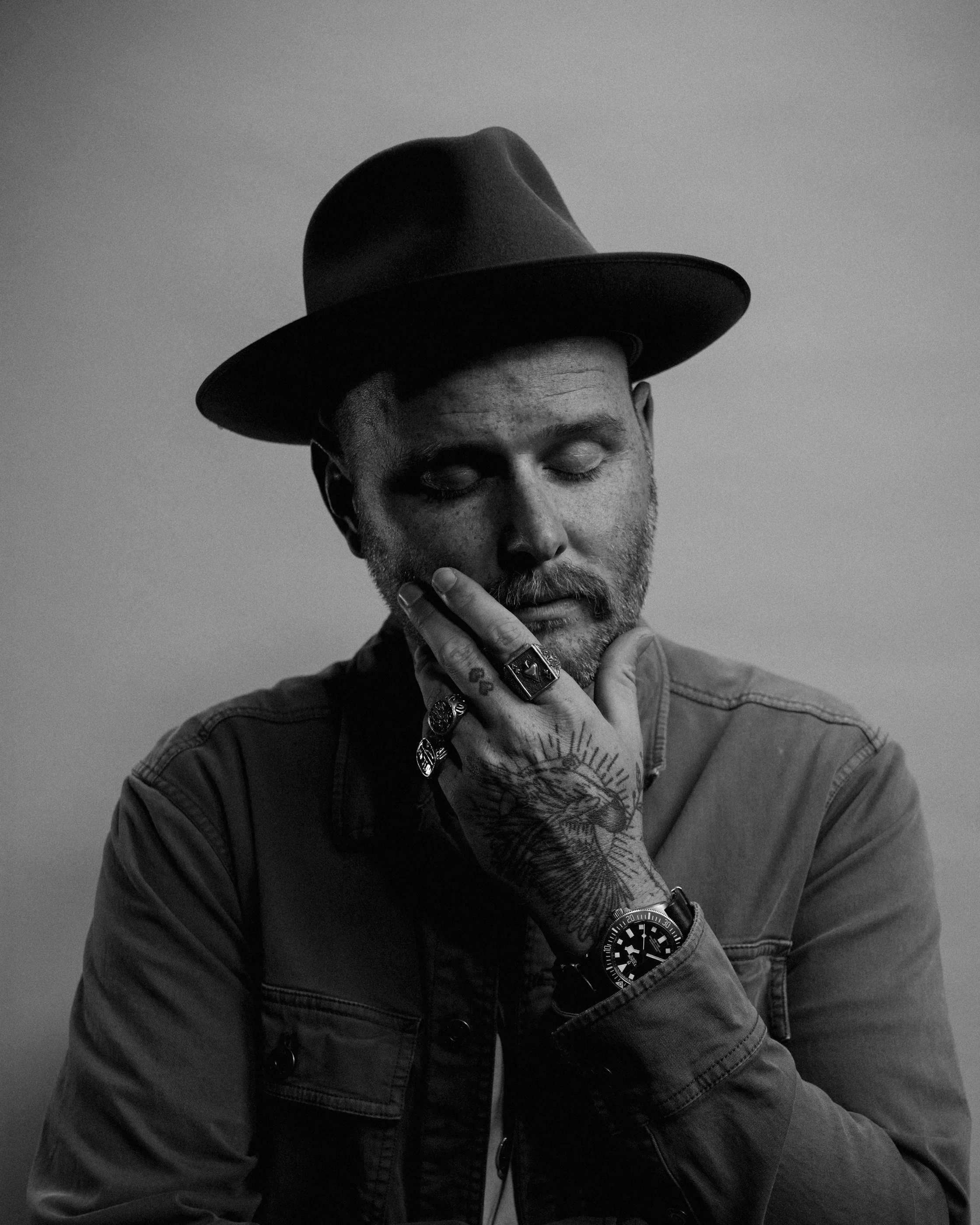 Black-and-white portrait of Dave Herring in a dark hat with his eyes closed and a tattooed hand against his chin.