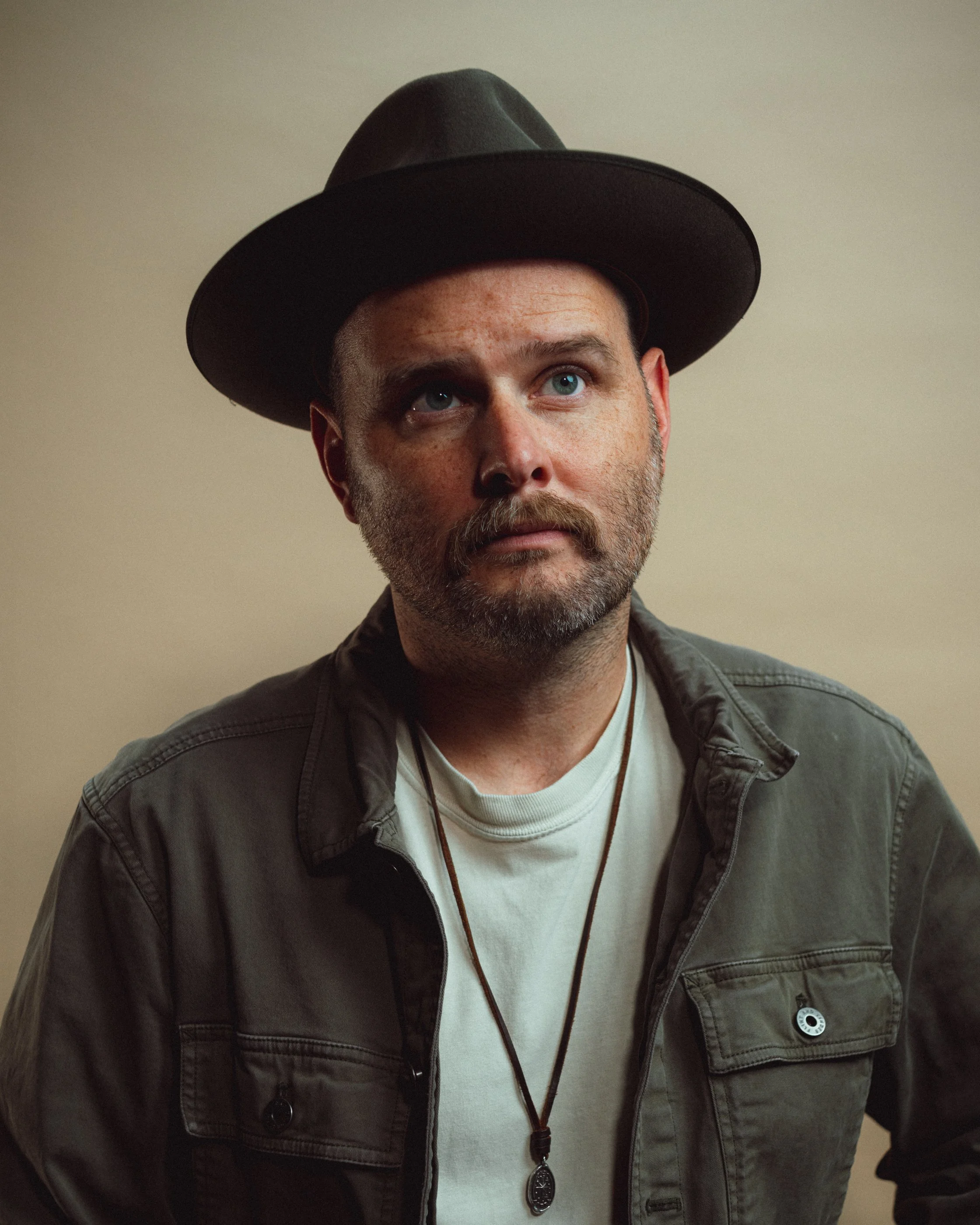 Dave Herring in a dark hat and olive overshirt looking up against a warm tan backdrop, with a pendant necklace.