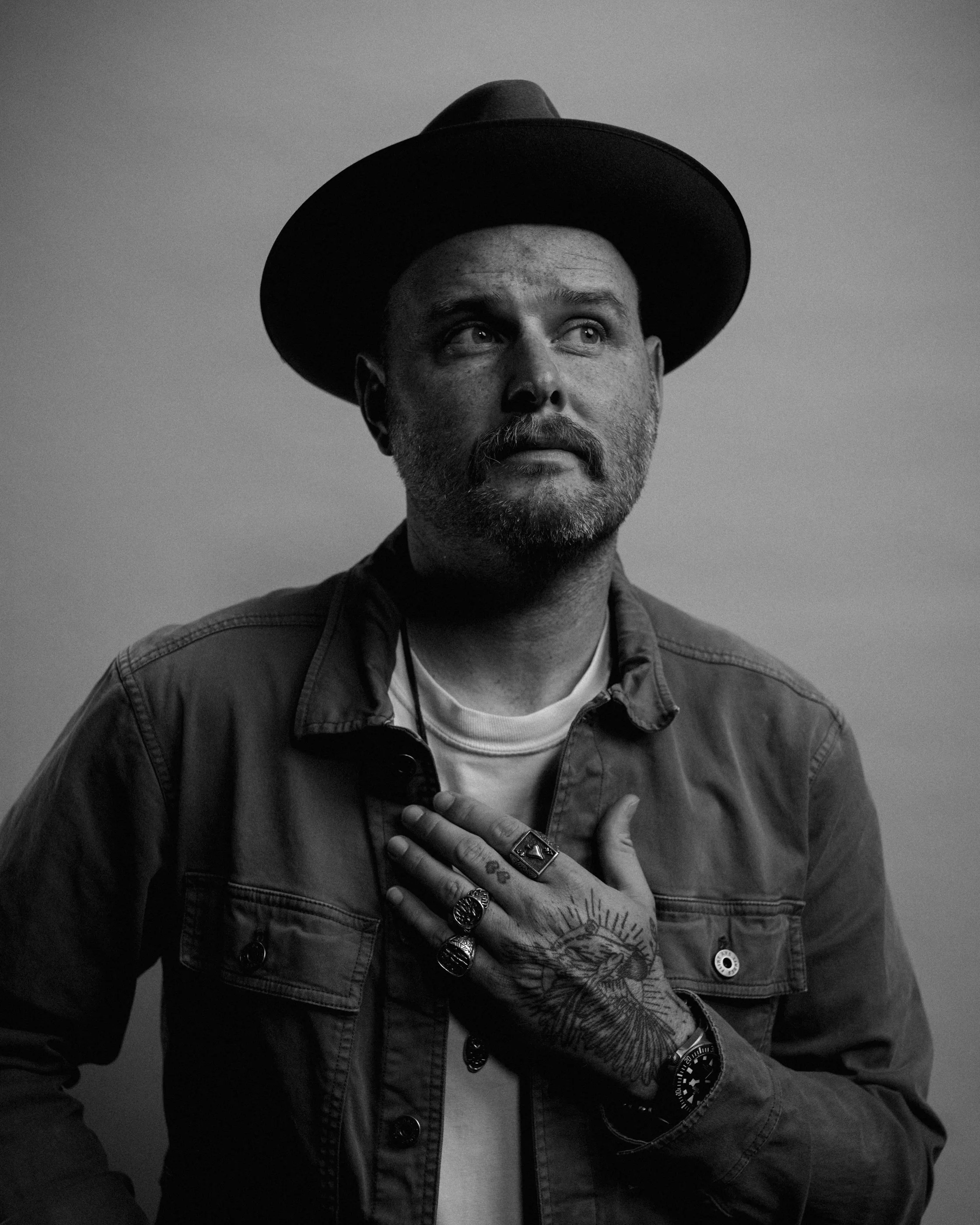 Black-and-white portrait of Dave Herring in a dark hat and overshirt looking up, one tattooed hand on his chest.