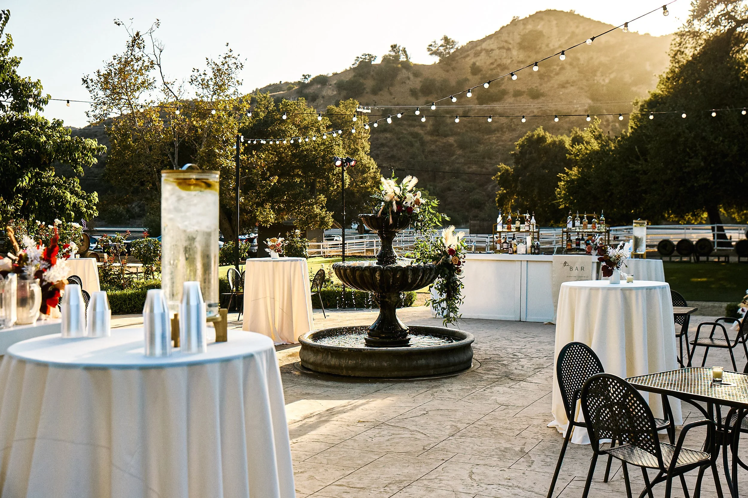 Outdoor event setup with round tables, some with floral centerpieces, a bar area with bottles, and a large fountain in the center, against a backdrop of mountains and trees, during sunset.