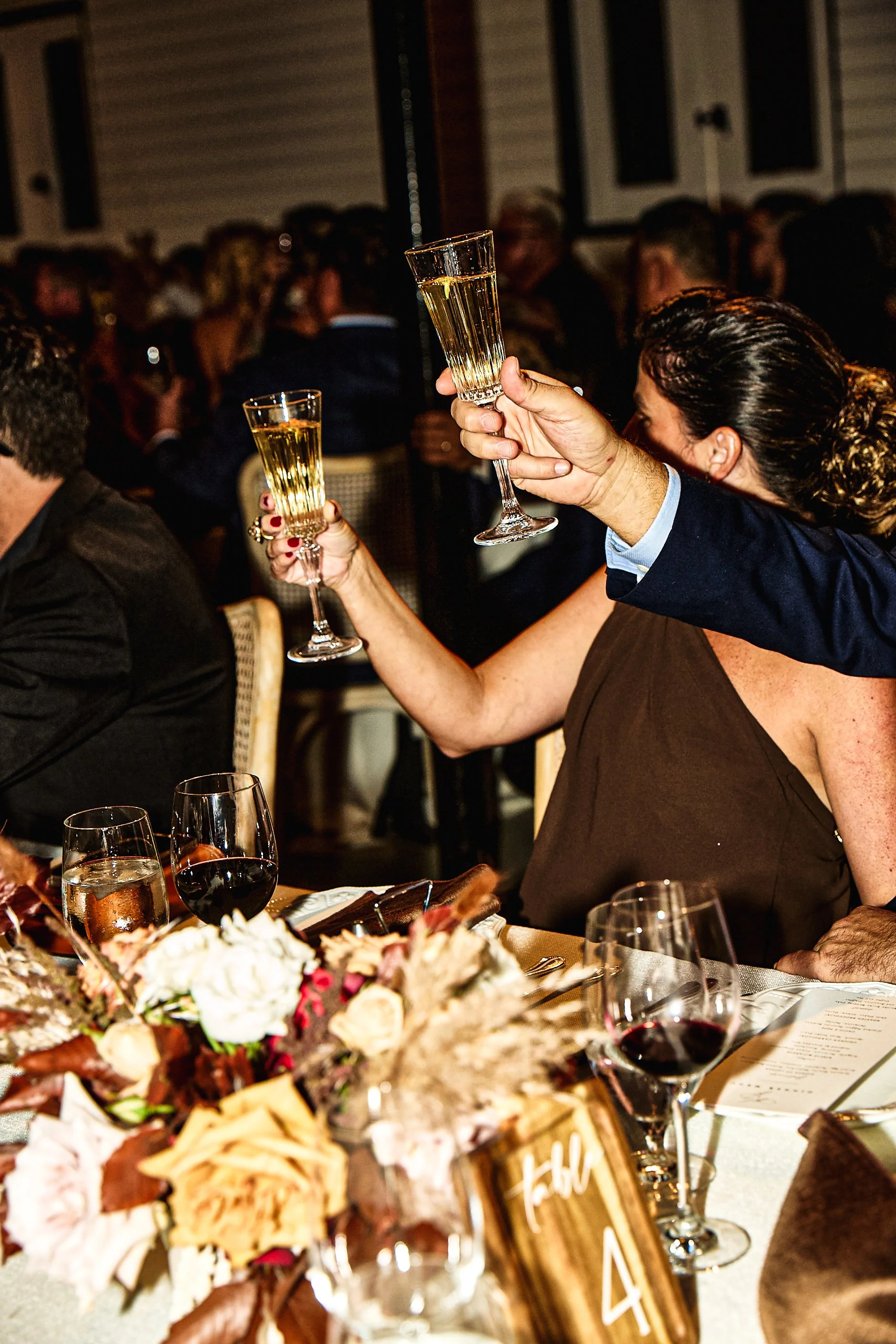 People at a banquet raising glasses of champagne for a toast. The table is decorated with flowers and glassware, and there are many guests in the background.