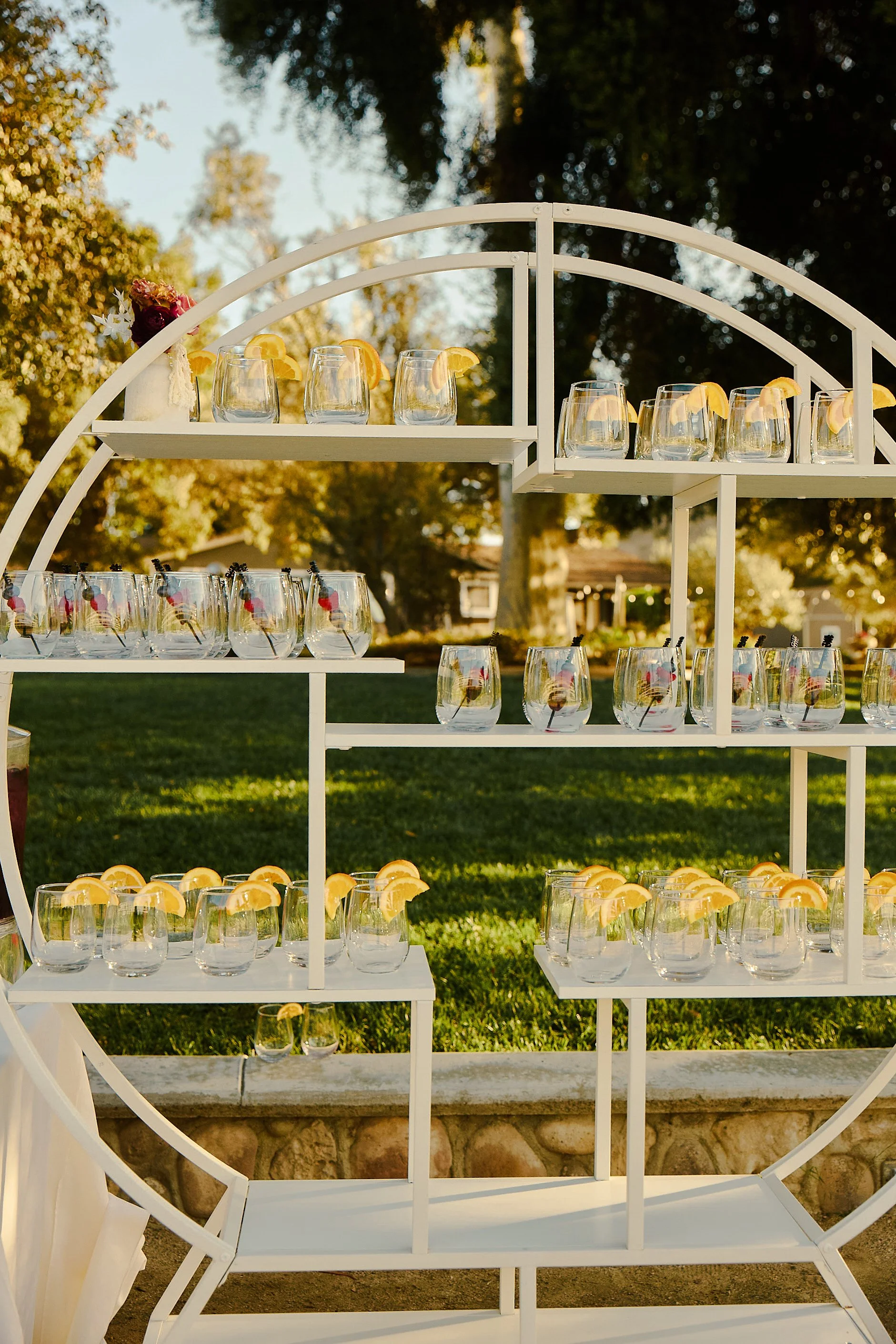 Display stand with glasses of water with lemon slices and cherries, set outdoors in a park with trees in the background.