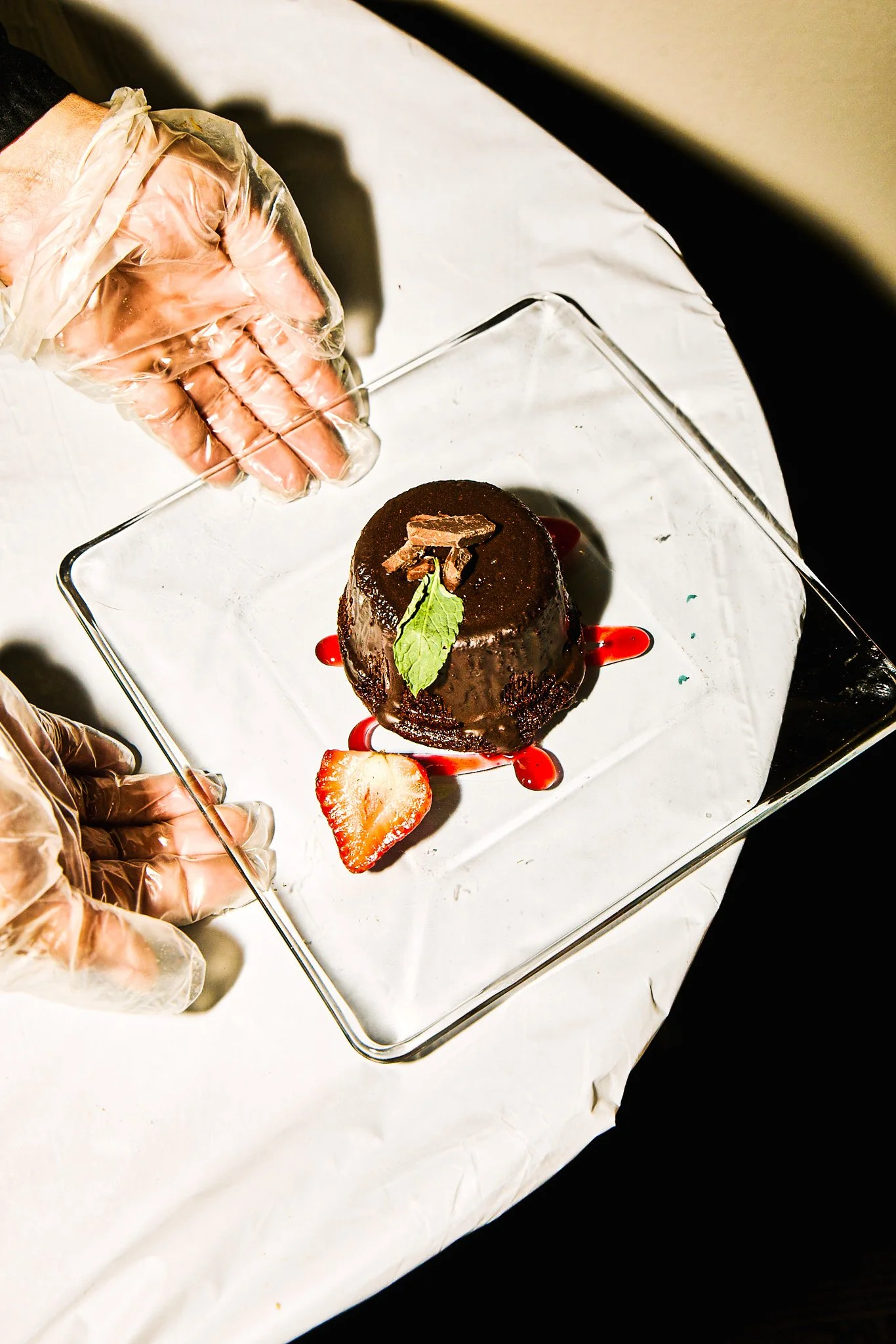 A person wearing transparent gloves presents a chocolate dessert on a glass plate, garnished with a mint leaf, a chocolate piece, a strawberry slice, and red sauce.