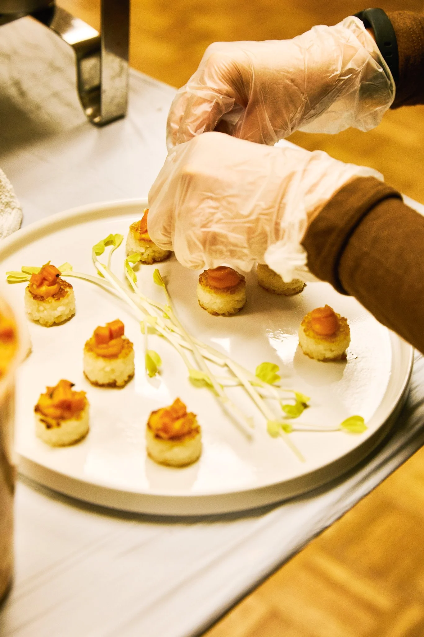 A person's hands, wearing gloves, preparing small sushi bites on a white tray, with some microgreens for garnish.