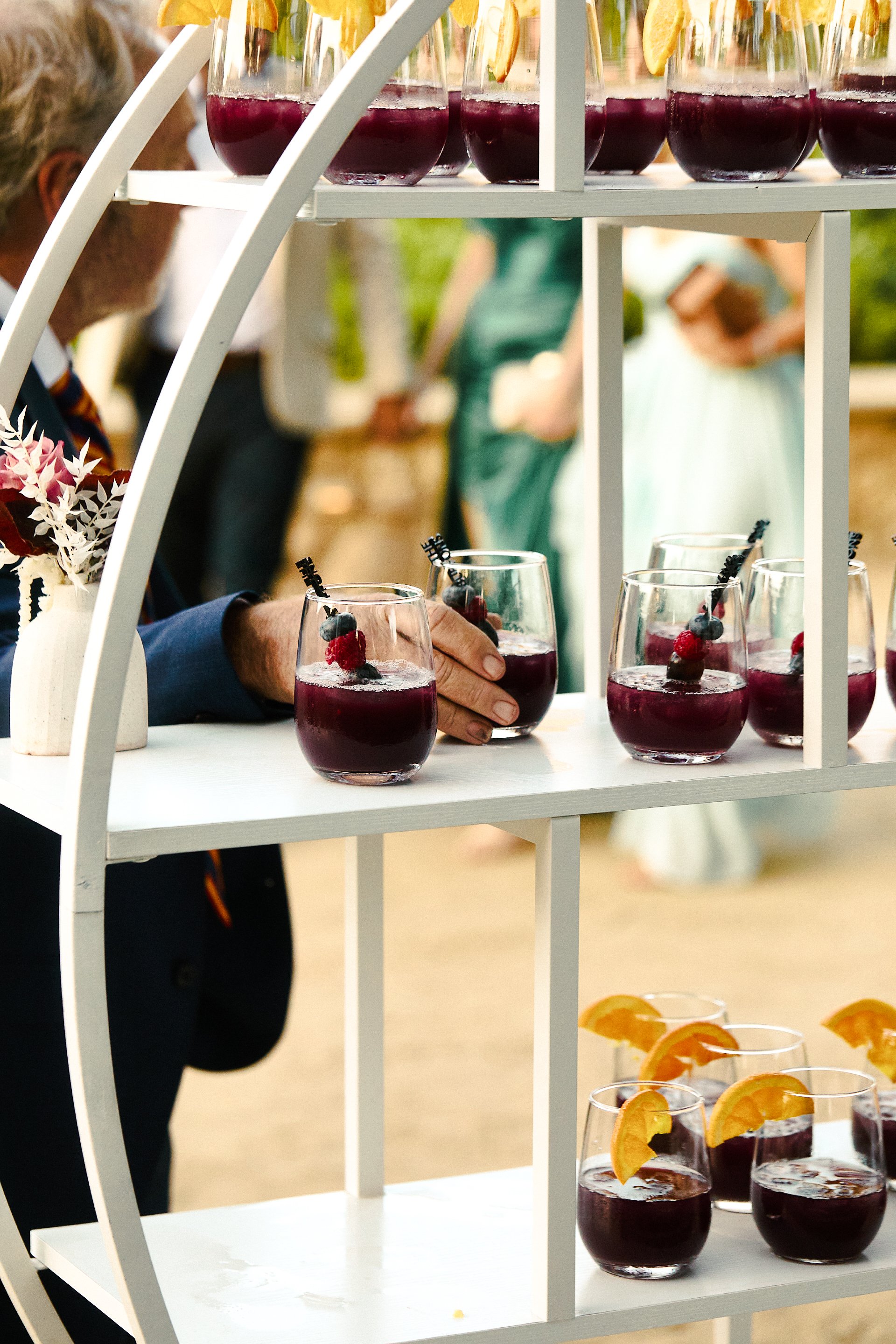 Glasses of dark red beverages garnished with berries and orange slices on a white shelf, with guests in the background at an outdoor event.