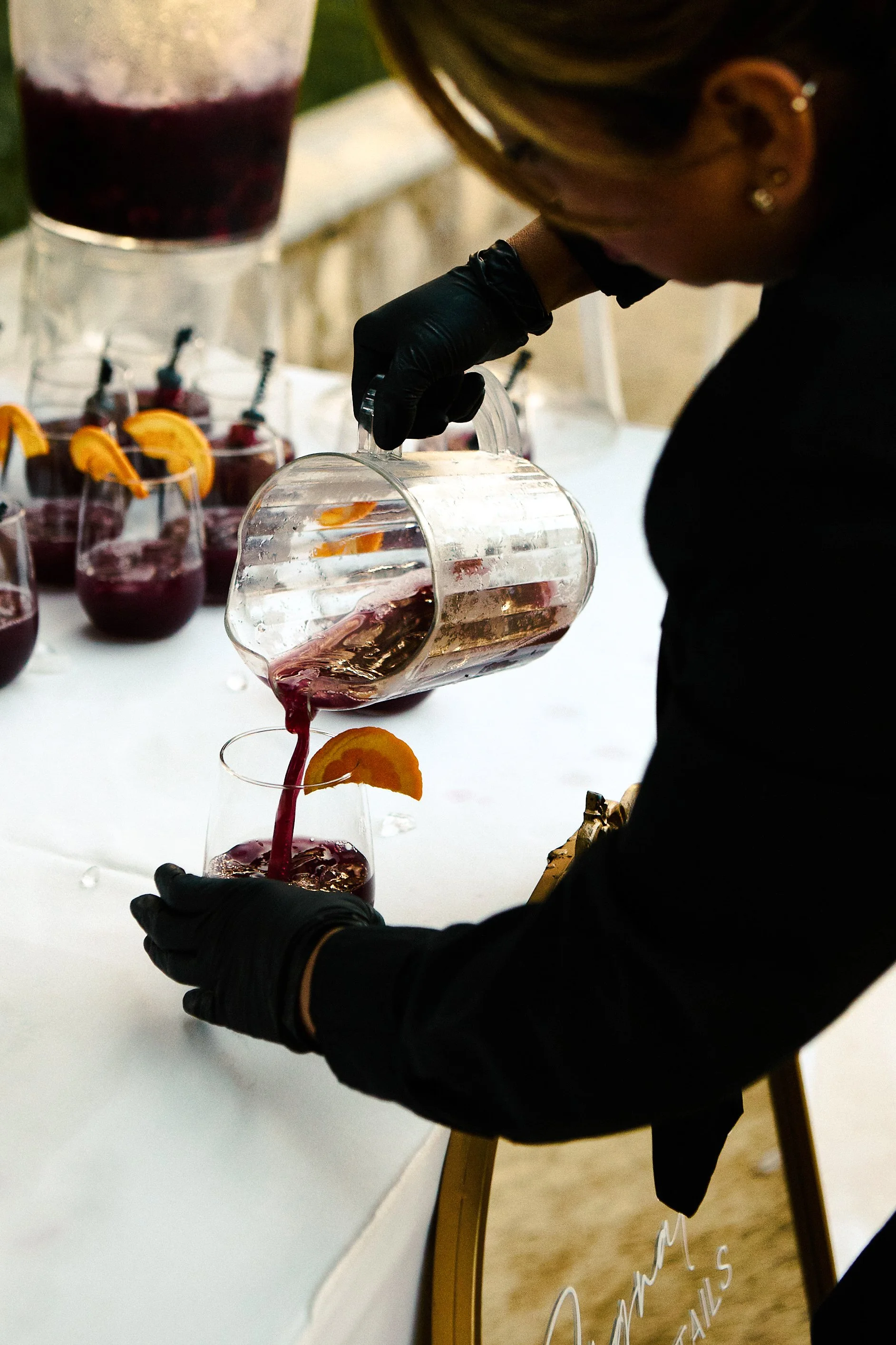 A person wearing black gloves pouring a dark purple beverage from a pitcher into a glass with an orange slice garnish.
