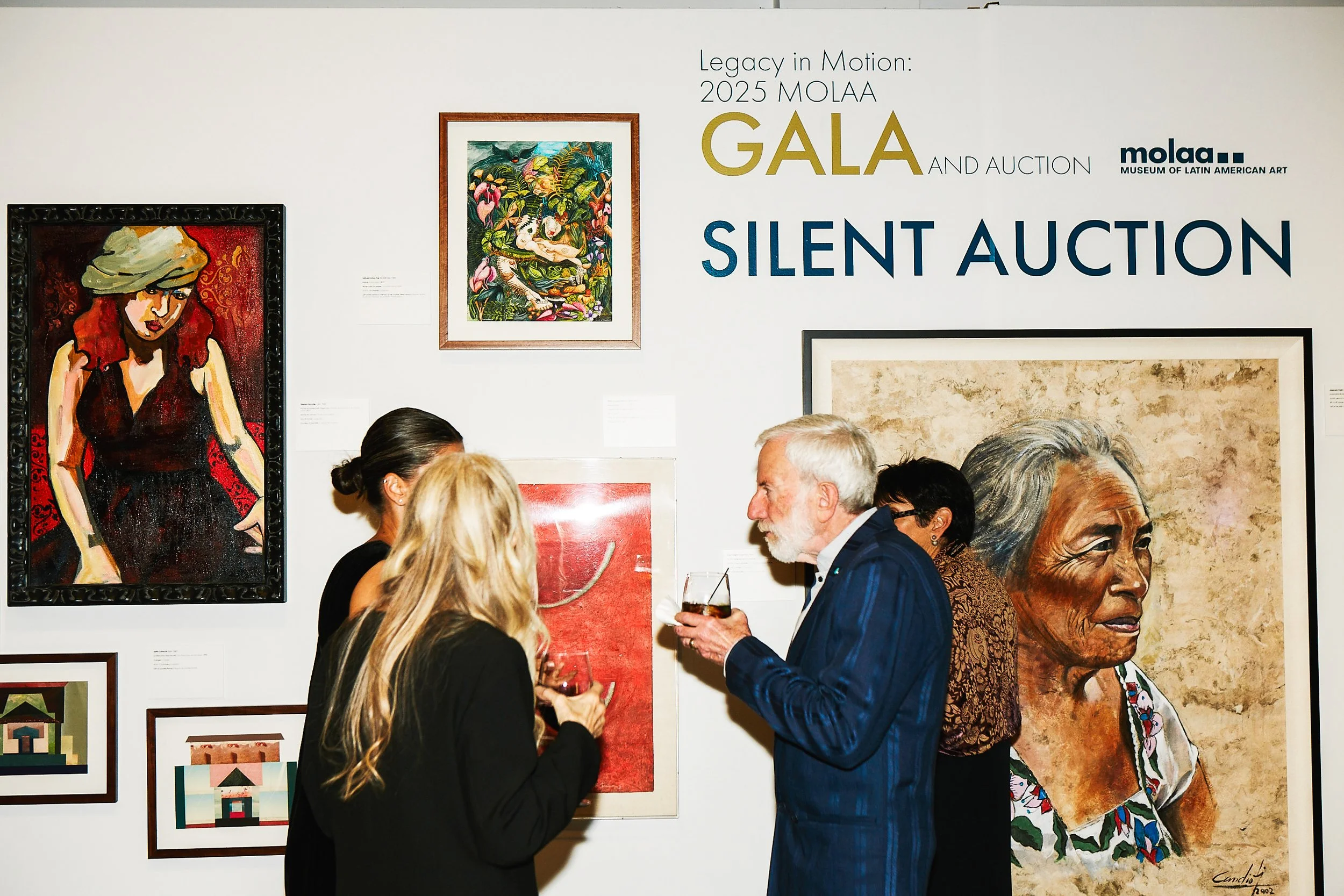 People attending a TGIS catered event in front of colorful paintings, including a portrait of a woman with gray hair and traditional clothing, with text indicating this is the 2025 MOLAA Gala and Silent Auction.