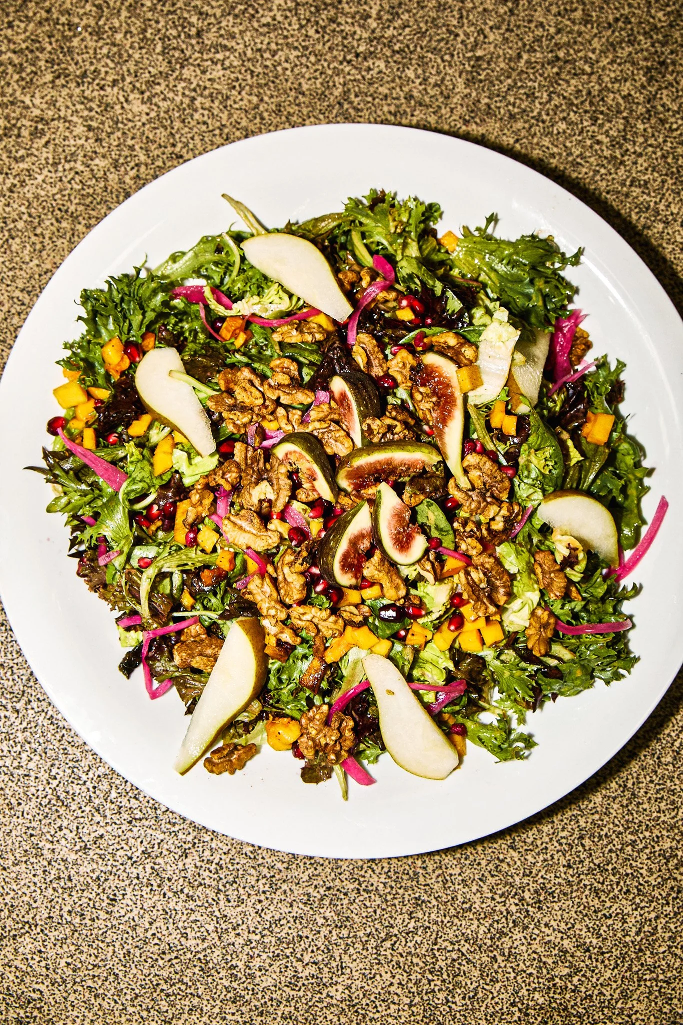 A white plate of mixed green salad with sliced pears, figs, walnuts, diced yellow bell peppers, and pink pomegranate seeds.