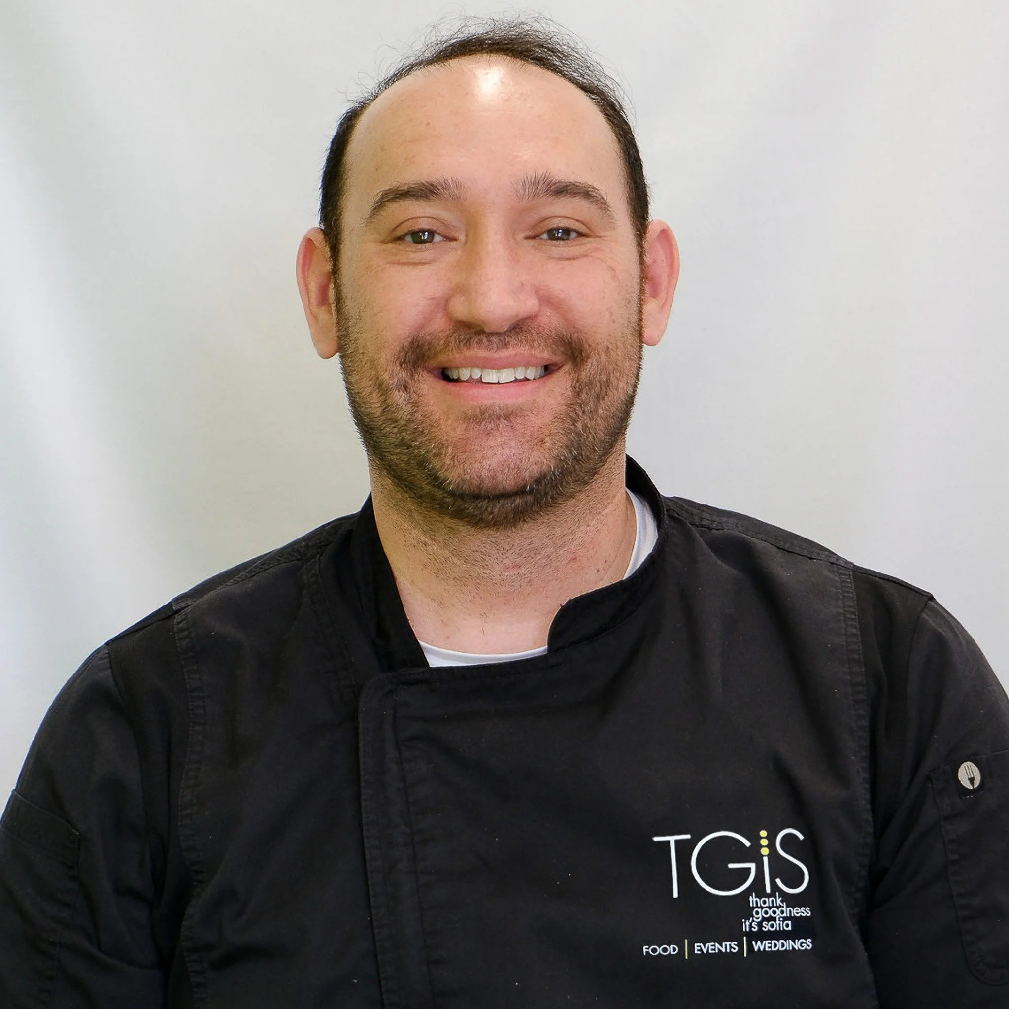 A smiling man with glasses and a beard, wearing a black chef jacket, standing against a gray background.