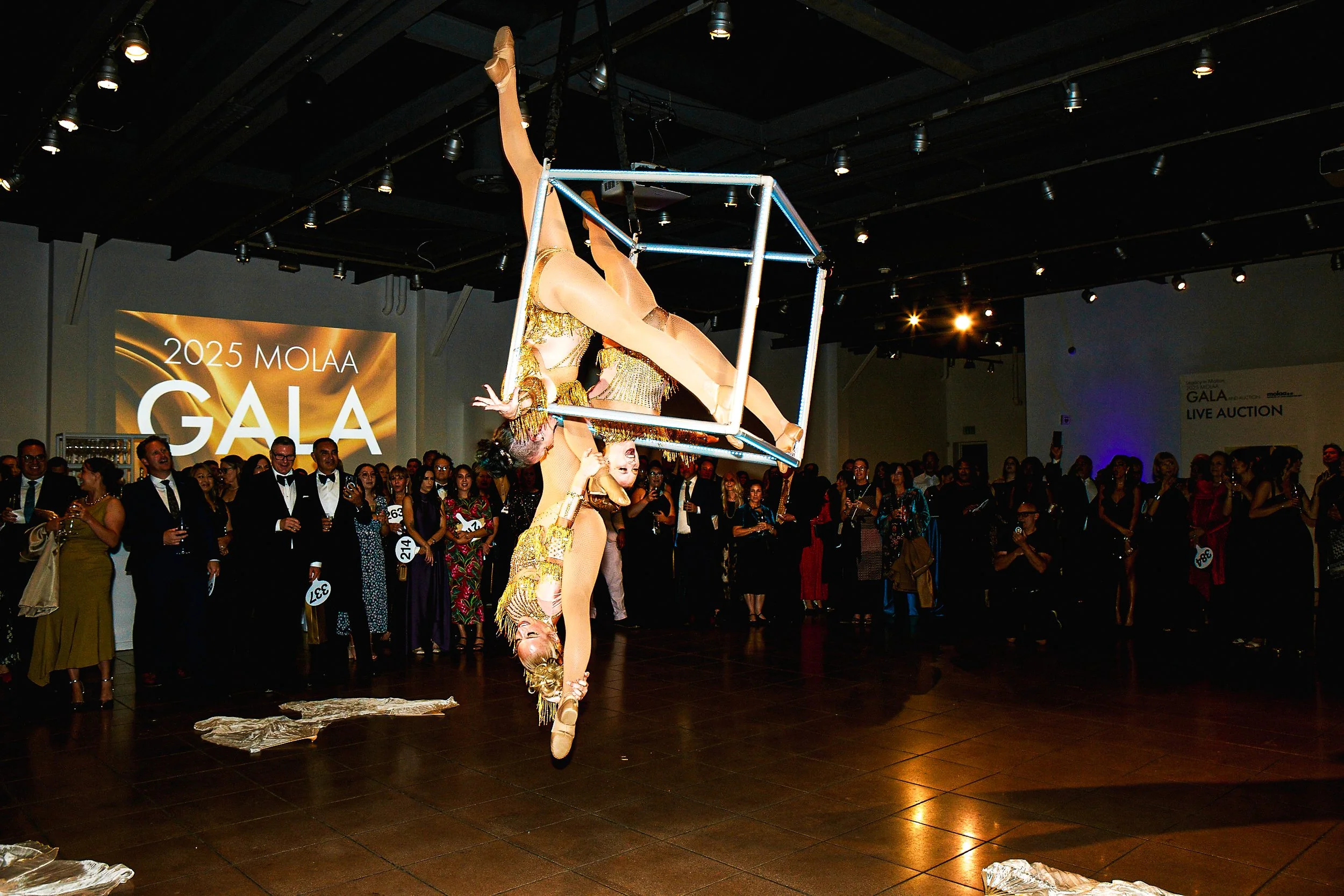 Two acrobats performing a balancing act with one balancing on the other's shoulders while holding a cube above their heads at the 2025 MOLAA Gala, with an audience watching in the background.