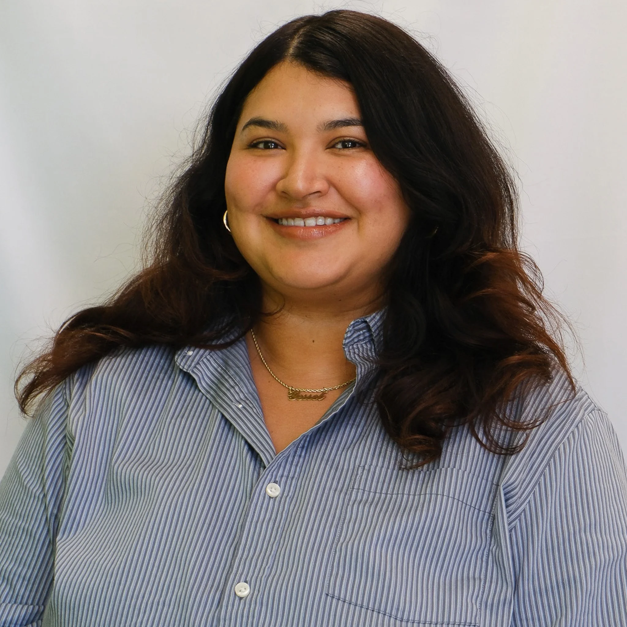 A smiling woman with long, dark brown hair and light makeup, wearing a black collared shirt, posing against a plain gray background.