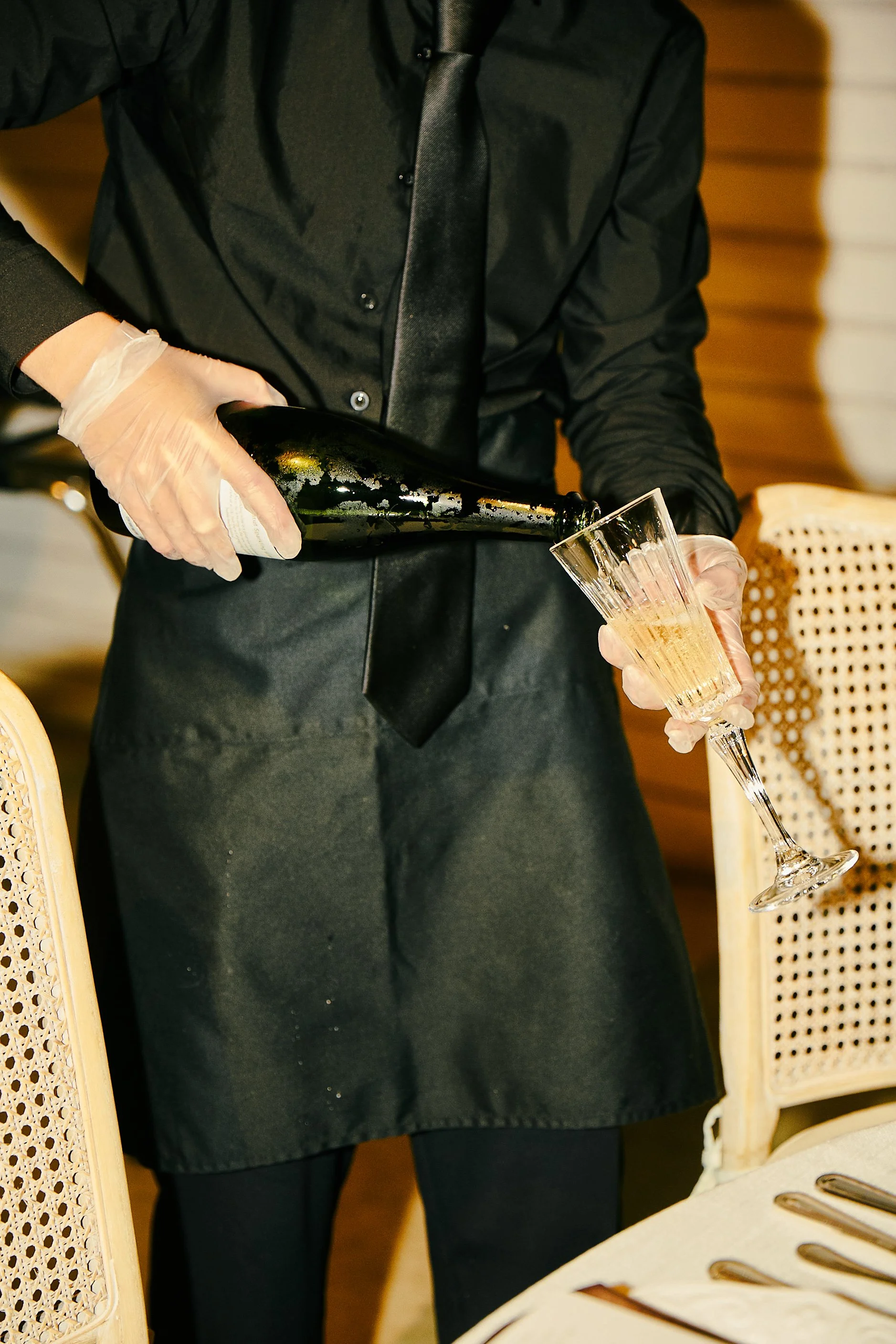 A person in black clothing and gloves is pouring champagne into a glass at a restaurant table.